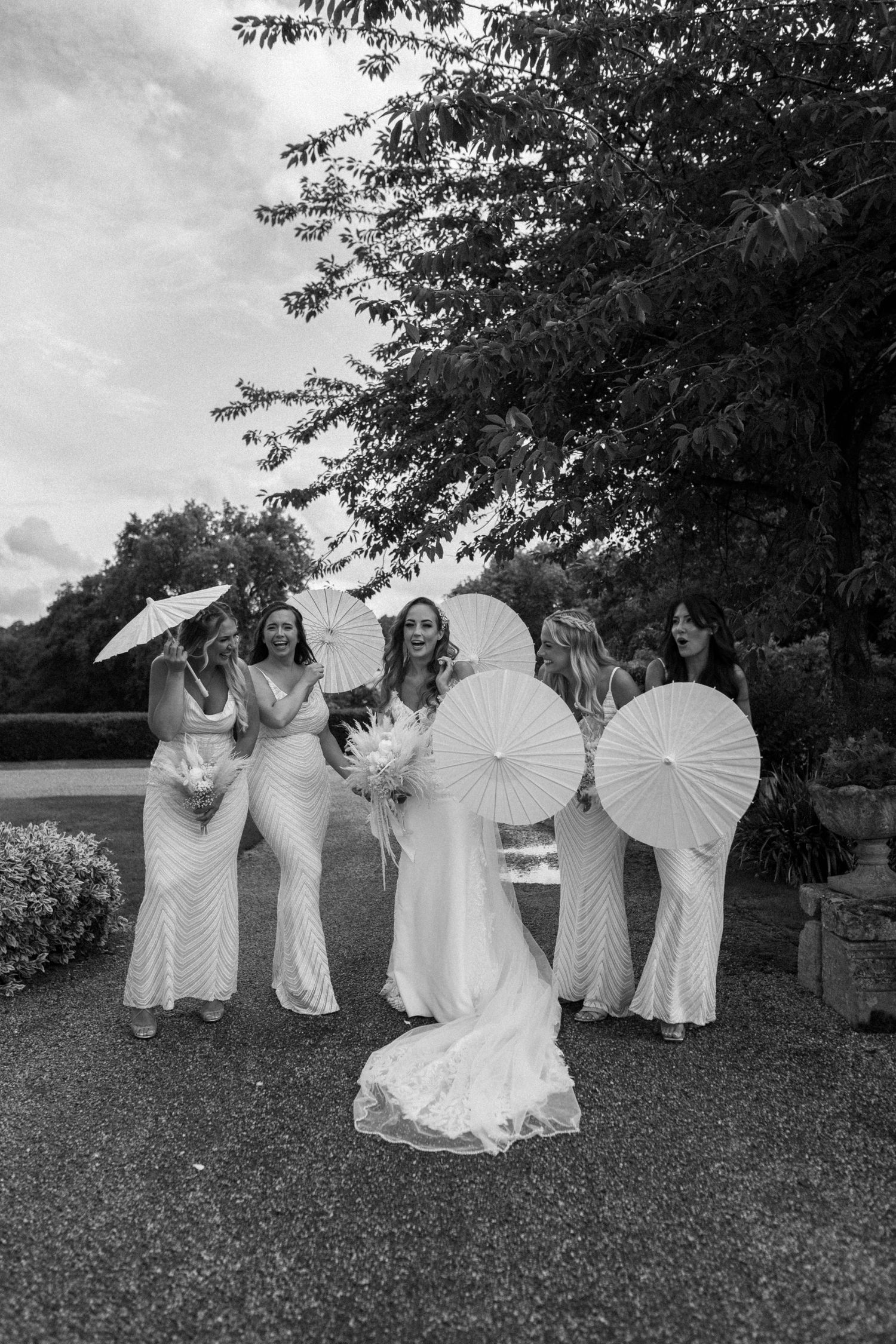 A bridal party posing with white parasols in a garden setting, captured in black and white.