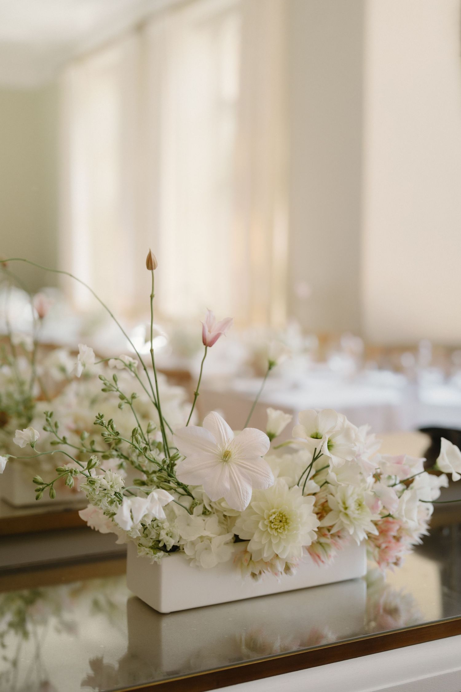 Elegant white rectangular vase with soft pink and white floral arrangement on mirrored table at wedding reception.