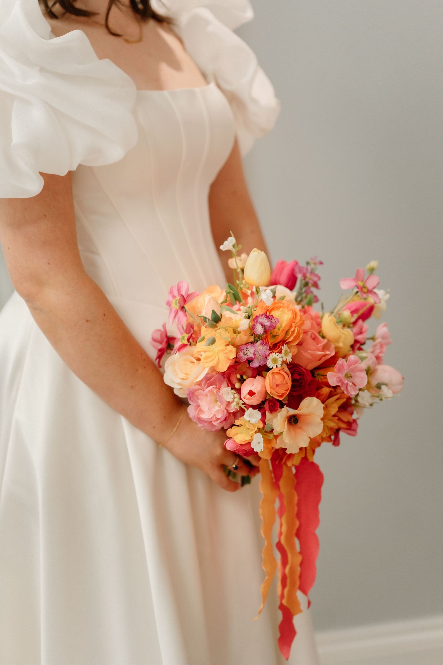 Bride in white puff-sleeve gown holding vibrant pink, orange, and yellow wedding bouquet with flowing ribbons.