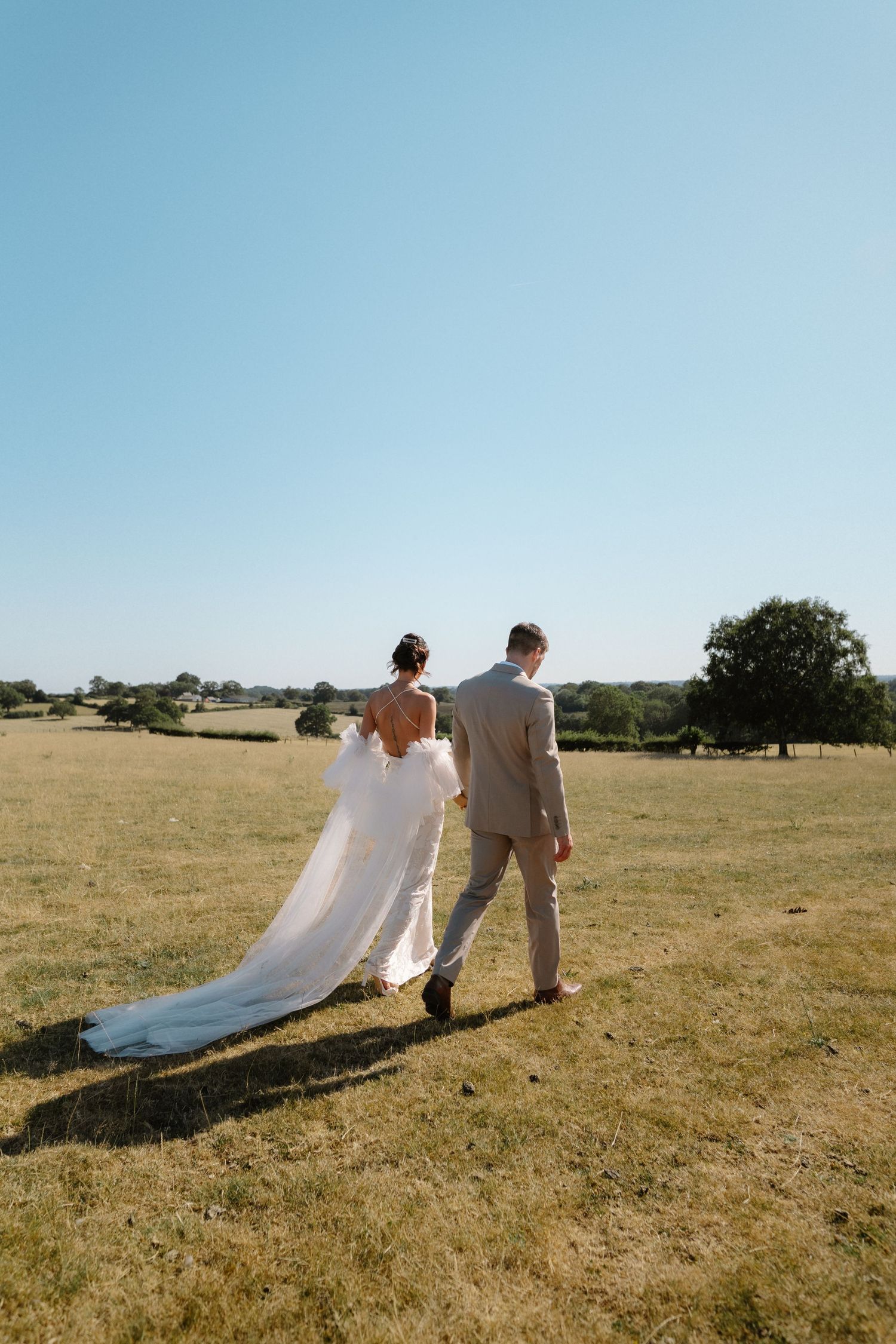 Bride and groom walking hand in hand across a sunny green field, bride wearing a flowing white gown with long train.