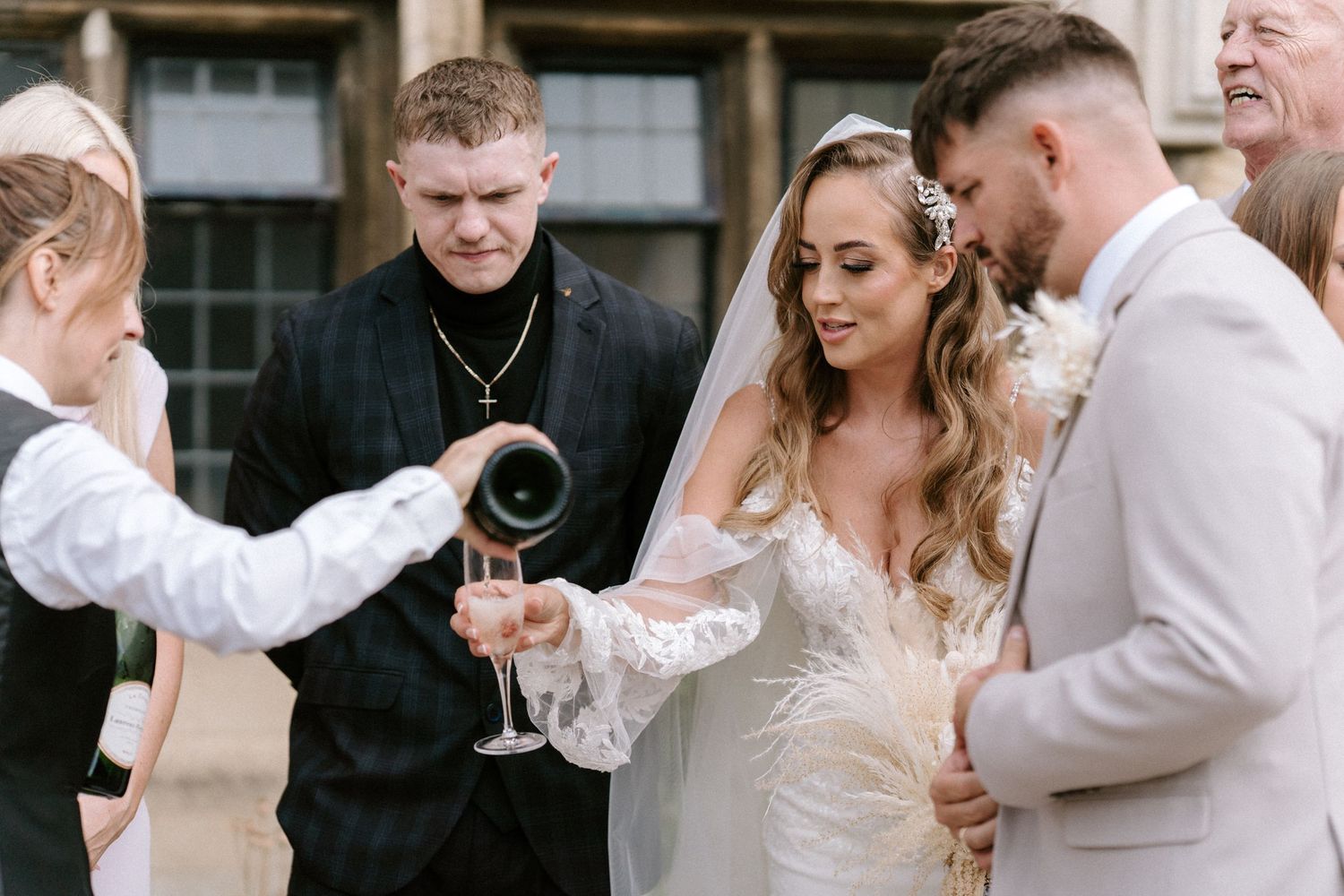 A couple in wedding attire shares a drink during an outdoor ceremony while others look on.