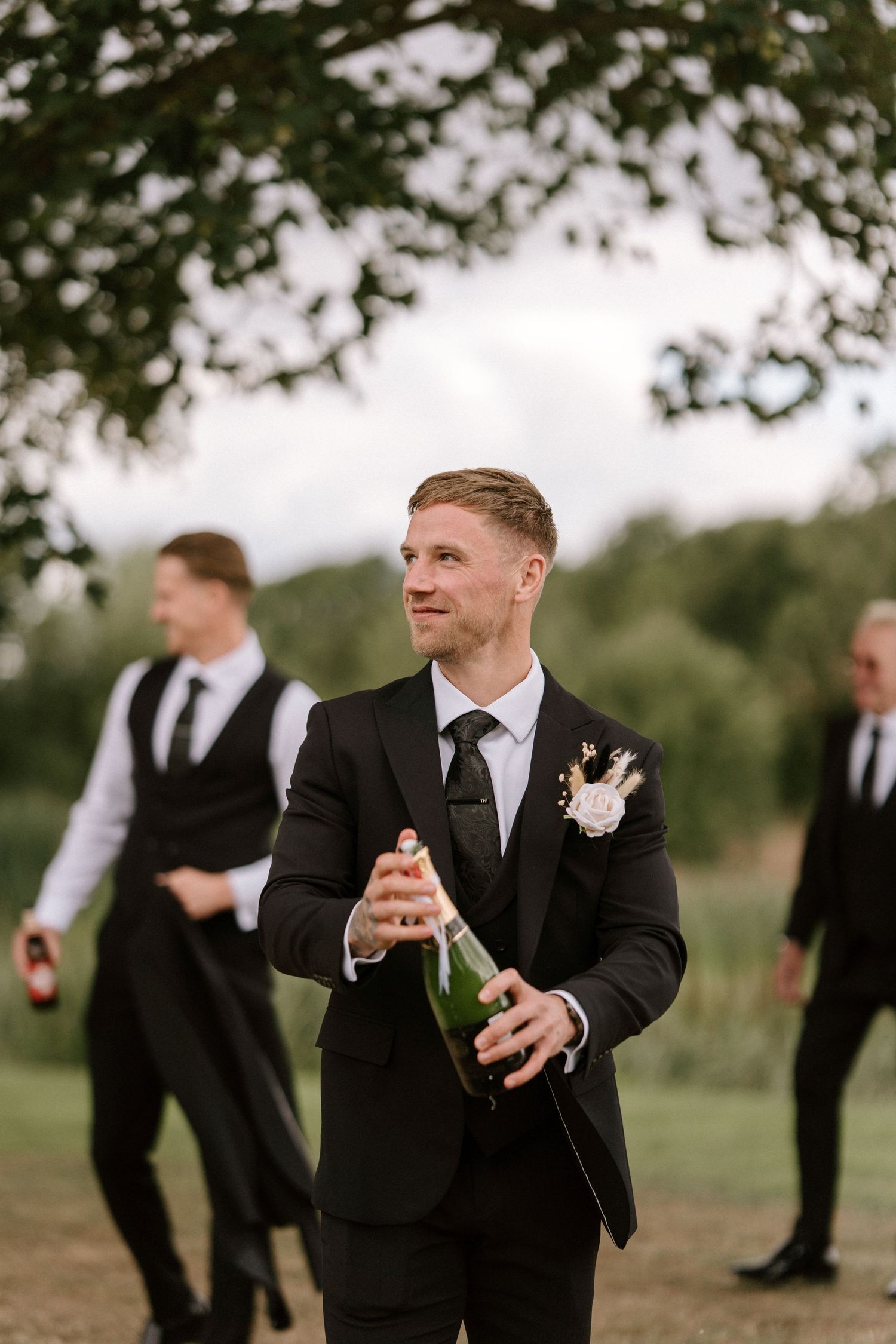 Outdoor wedding party member in black suit with boutonniere opening champagne under tree branches.