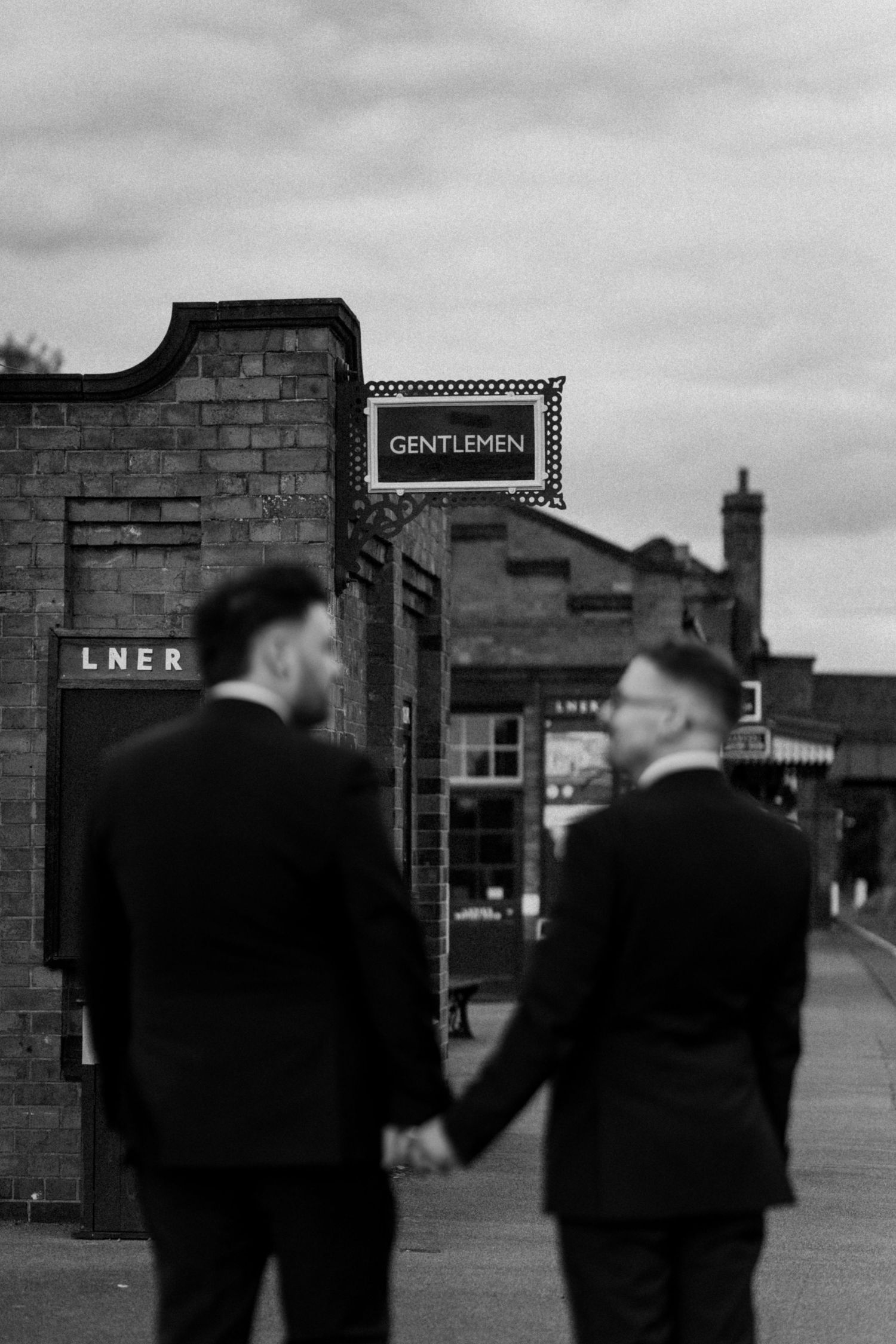 Two figures in suits holding hands near a vintage railway station with 'Gentlemen' sign at The Manor House at Quorn.