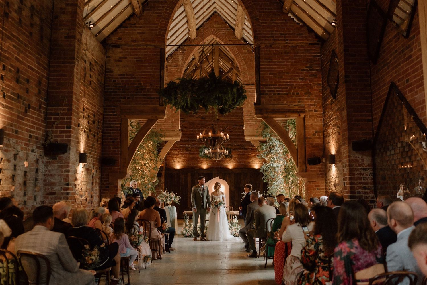 Wedding ceremony at the stunning brick barn venue of Shustoke Barn with vaulted ceilings and dramatic lighting.