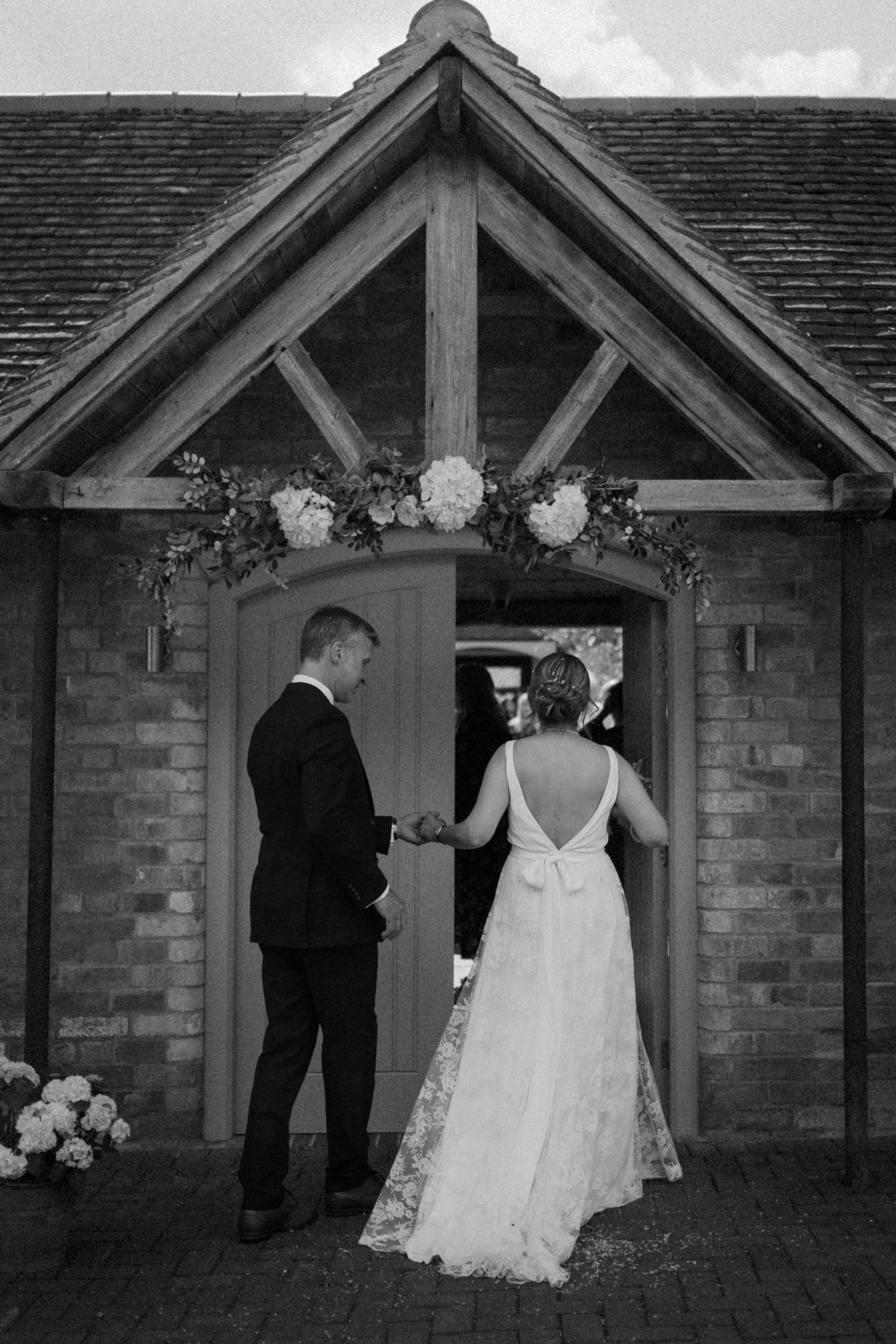 A couple stands in the doorway of a rustic brick building with floral decorations above during a wedding ceremony at Swallows Nest Barn.