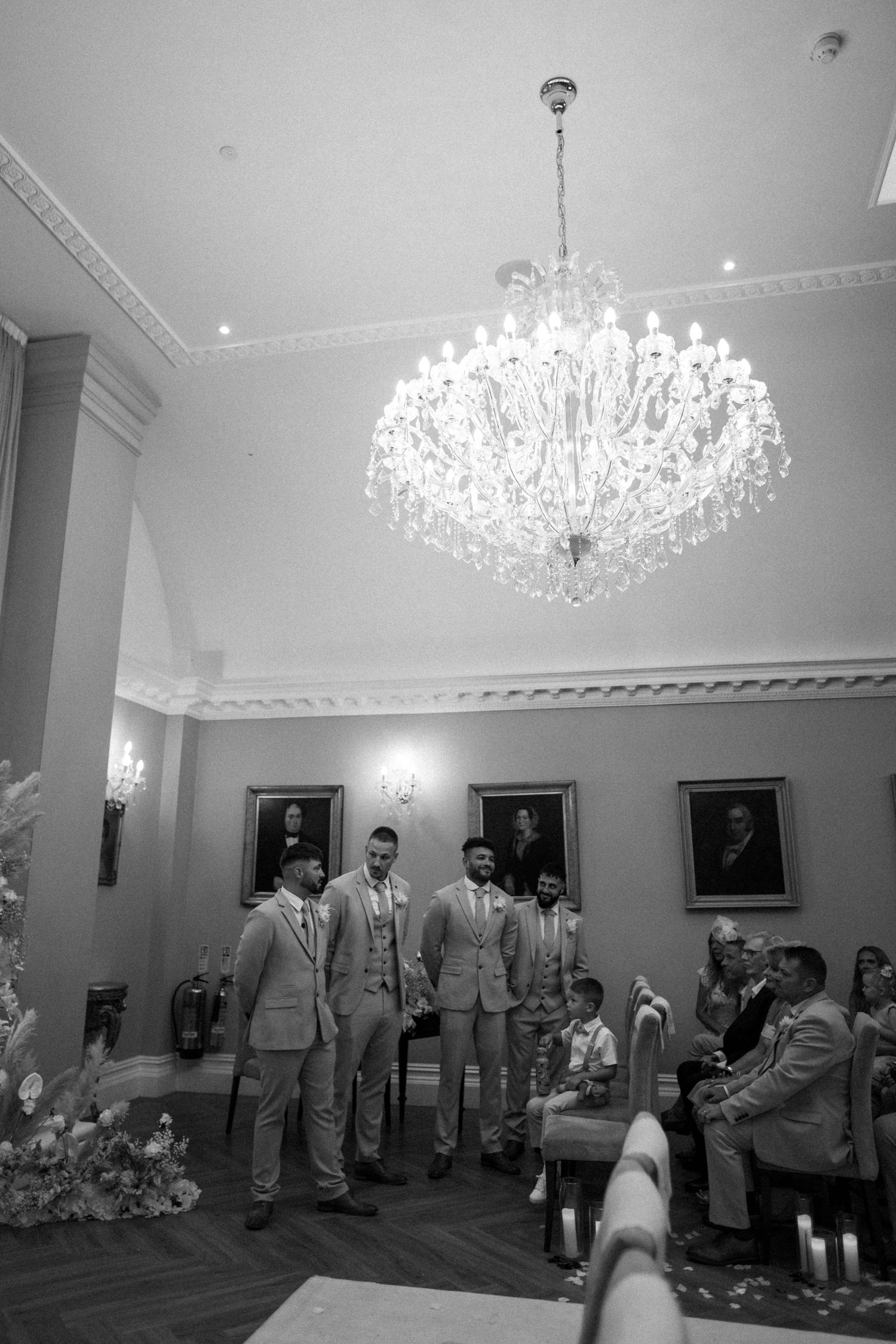 Elegant ballroom with crystal chandelier and groomsmen in light suits during wedding ceremony.