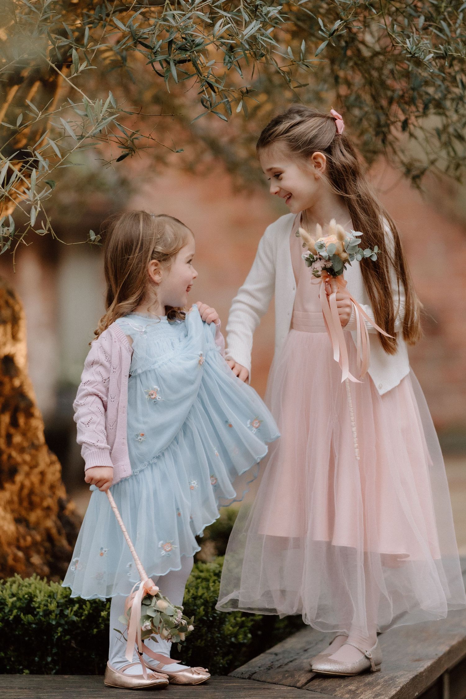 Two children in flowing dresses, one pink and one blue, share a sweet moment together under foliage at Shustoke Barn.