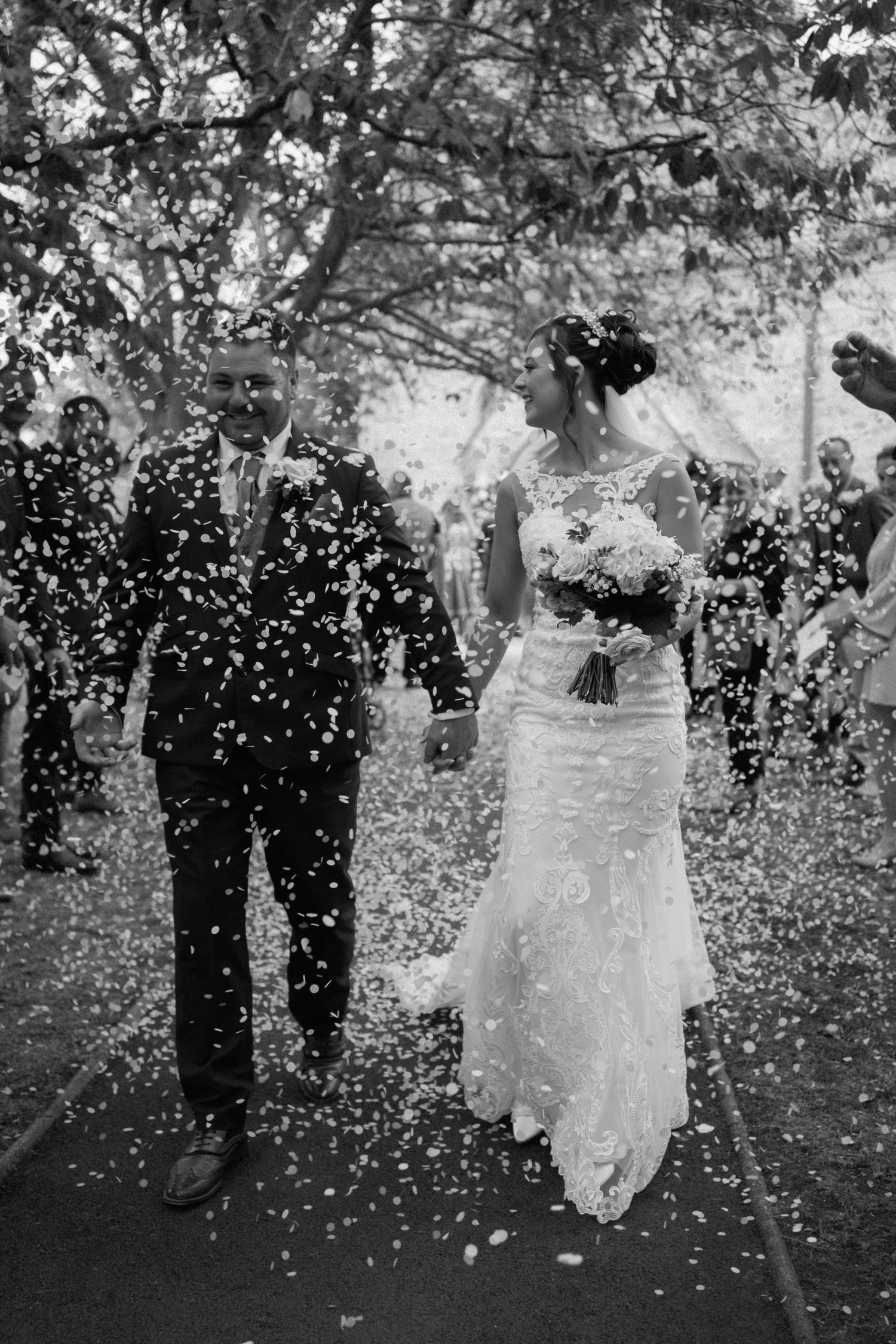 Black and white wedding photo showing couple walking through falling confetti in an outdoor garden setting.
