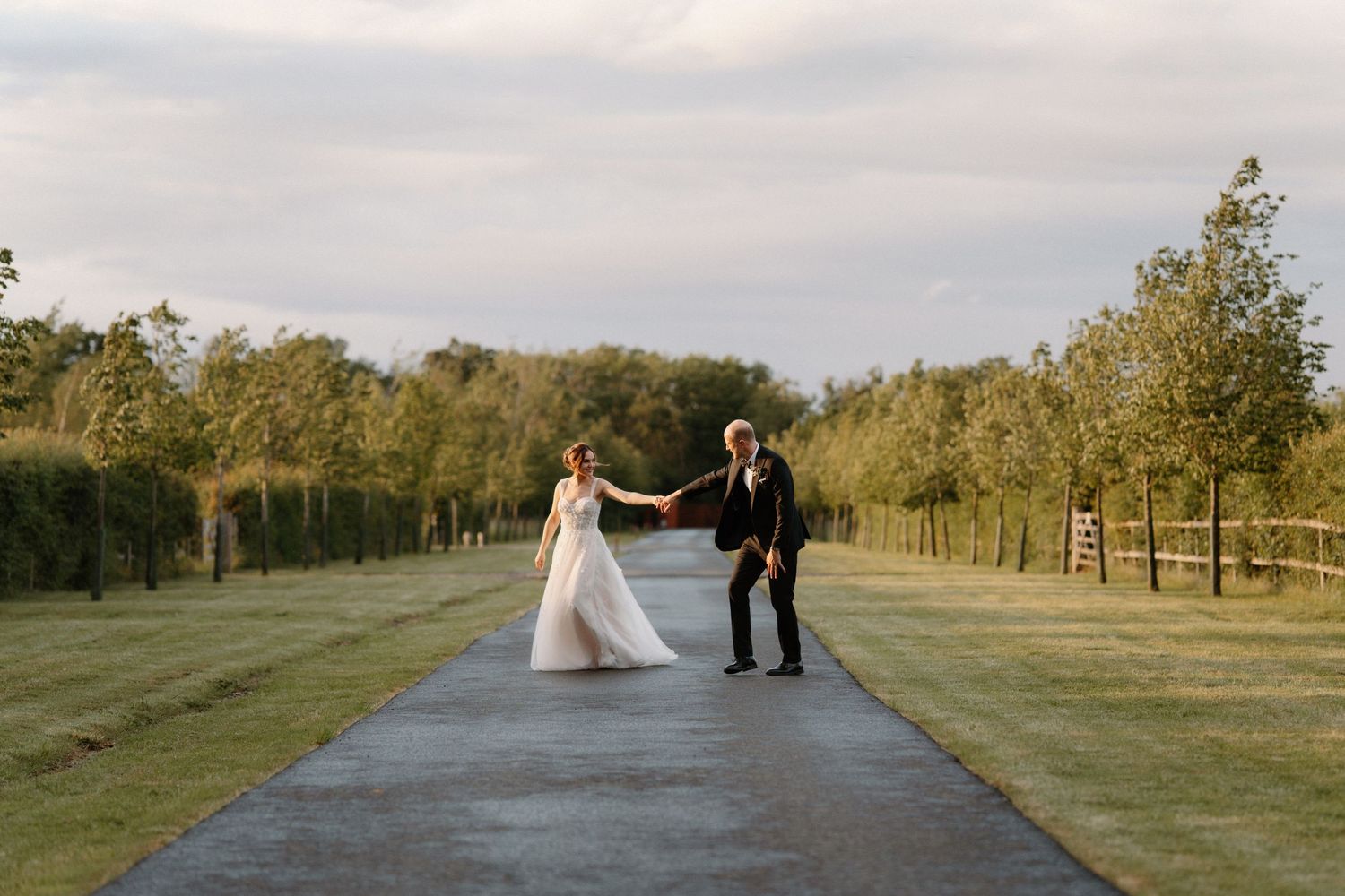 Bride and groom hold hands on a tree-lined path at golden hour during an outdoor wedding celebration.