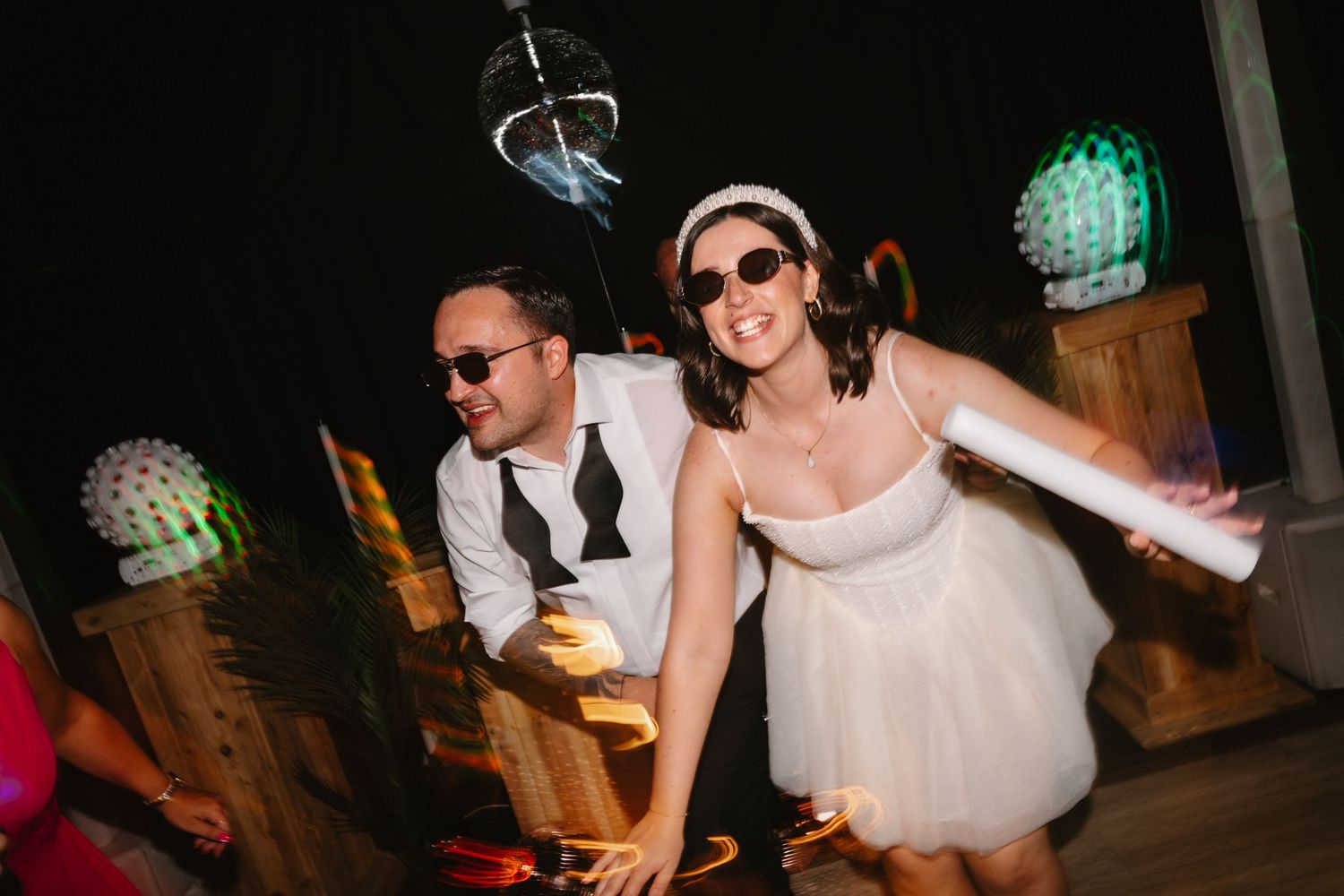 Bride and groom dancing joyfully at wedding reception, wearing sunglasses under disco ball lights at night.
