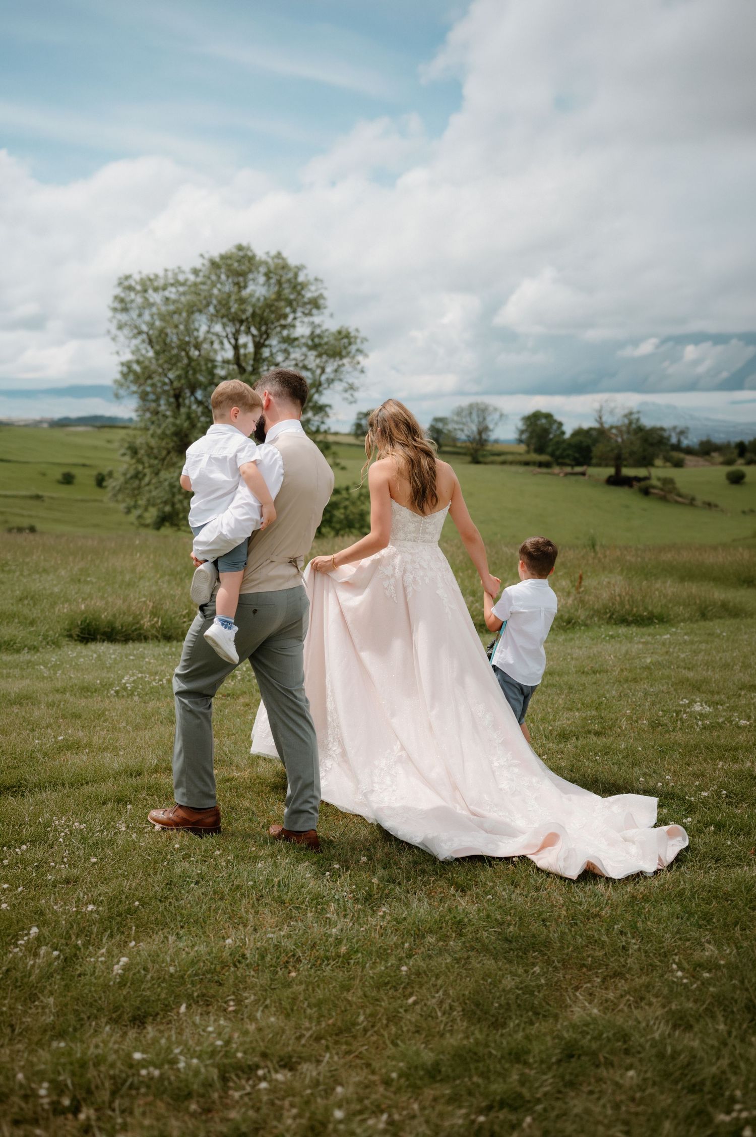 A bride in a white gown holds hands with her son while her groom carries a toddler across a lush green field.