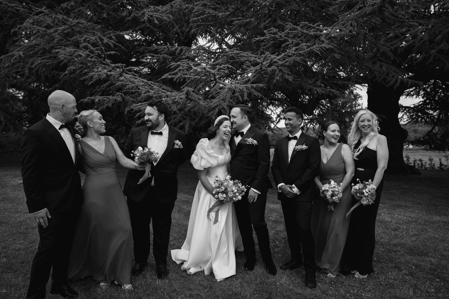 Black and white wedding party photo with bride, groom, and six attendants laughing outdoors under large trees.