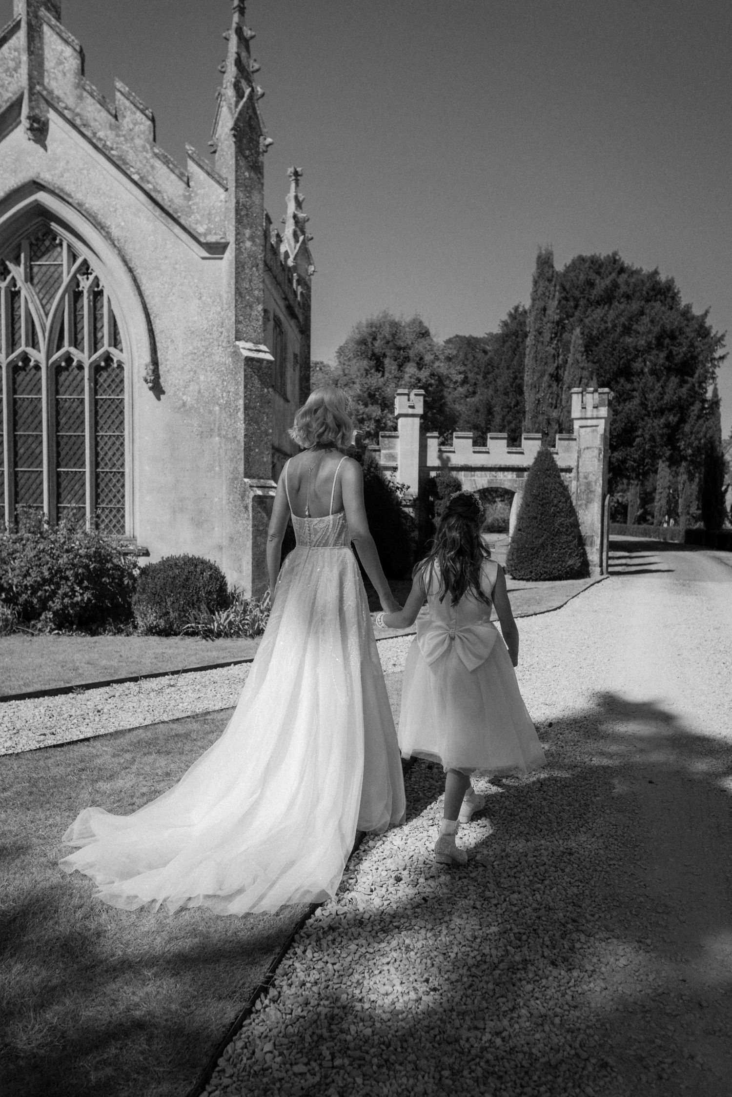 Bride in flowing tulle gown holds hands with flower girl outside a Gothic stone chapel.