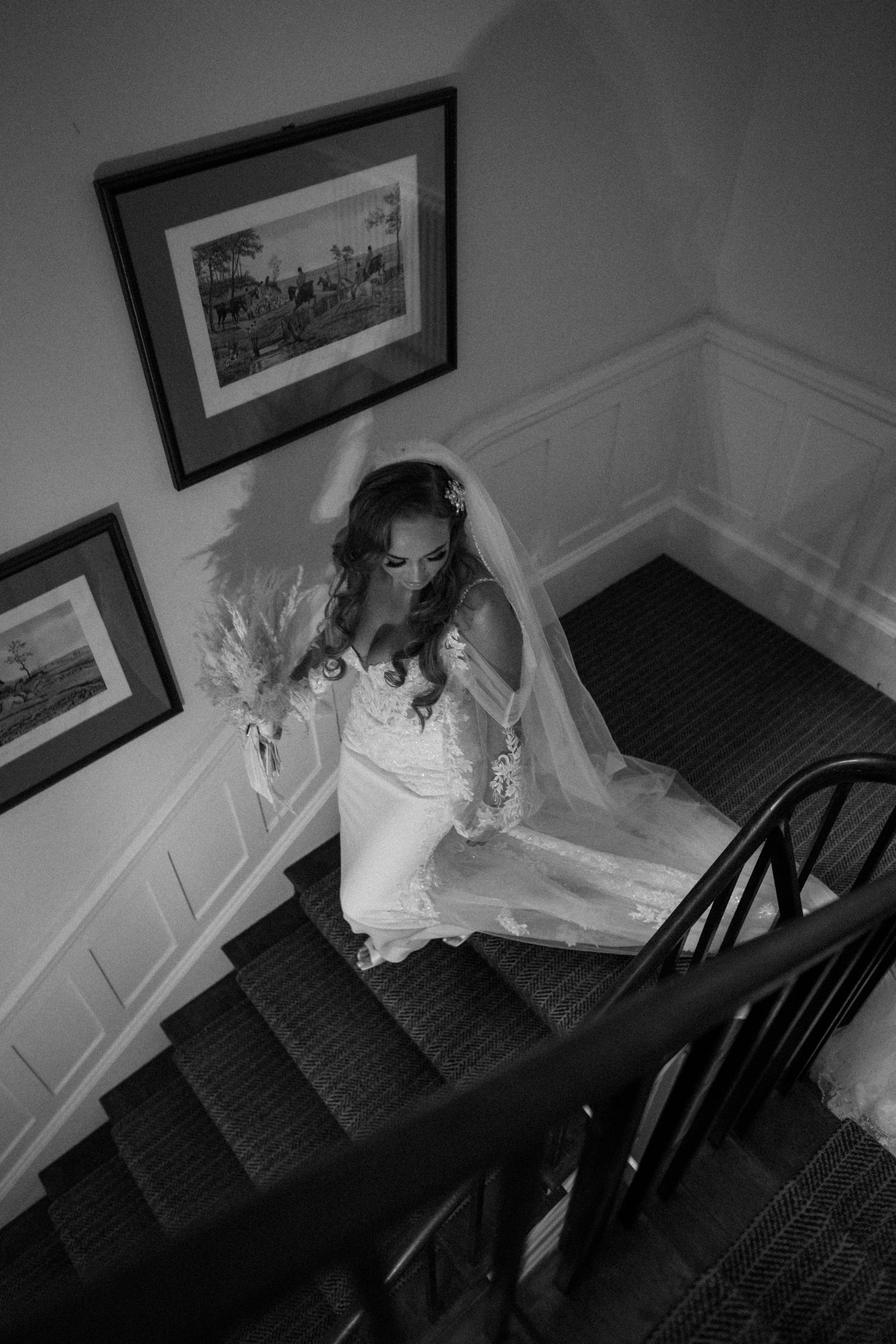 A bride in a white wedding dress descends a carpeted staircase with black railing and framed artwork on the walls.
