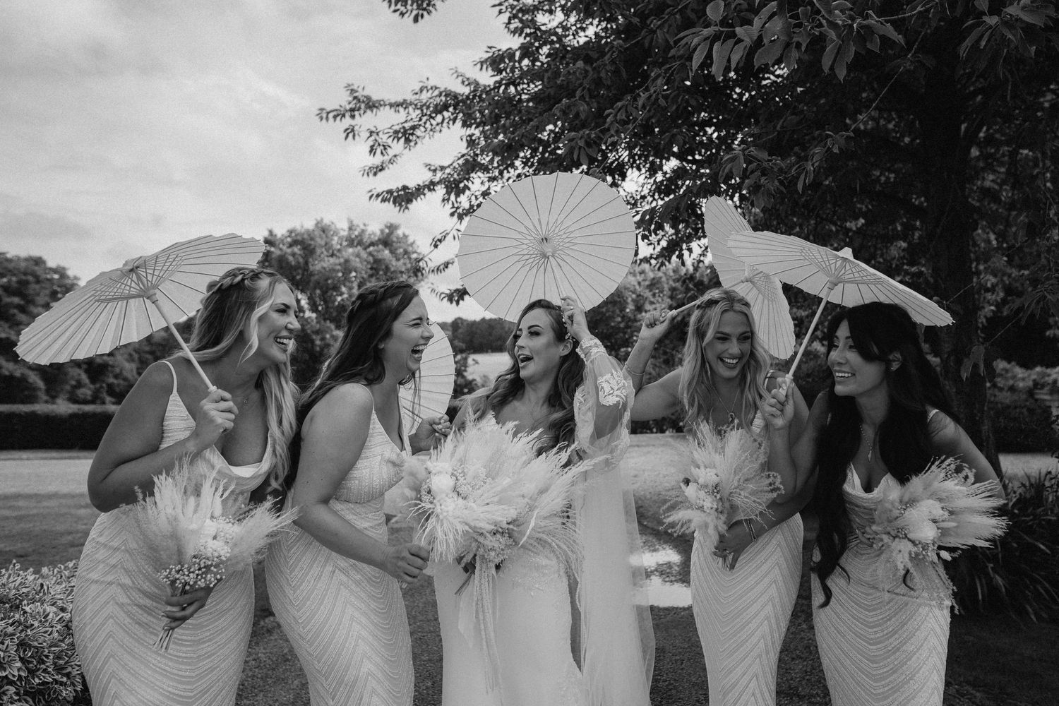 A bridal party carrying white parasols and bouquets poses together for a candid moment in black and white photography.