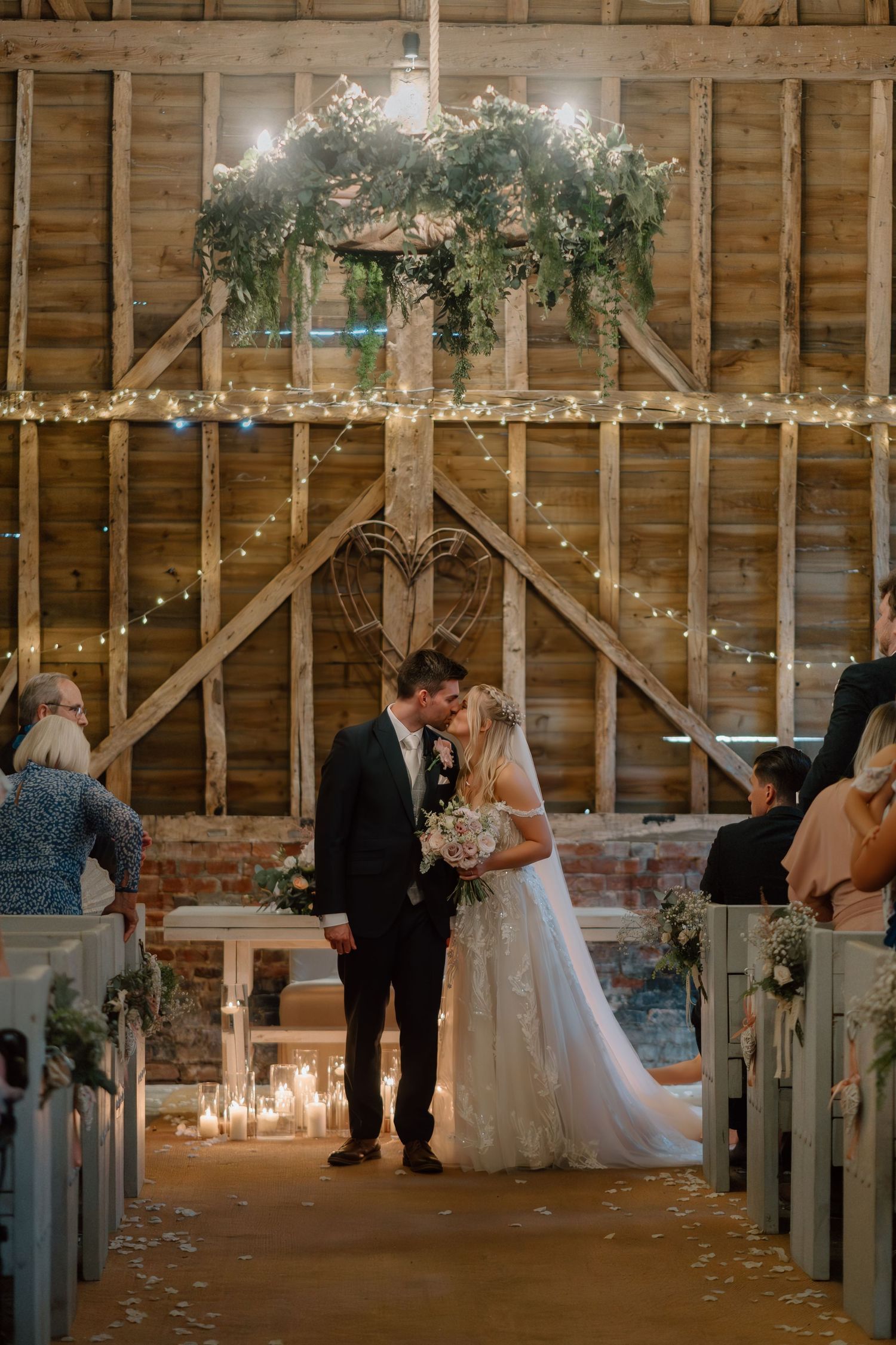 Rustic barn wedding ceremony with glowing string lights, floral arch, and candle-lined aisle in ethereal evening setting at Milling Barn.