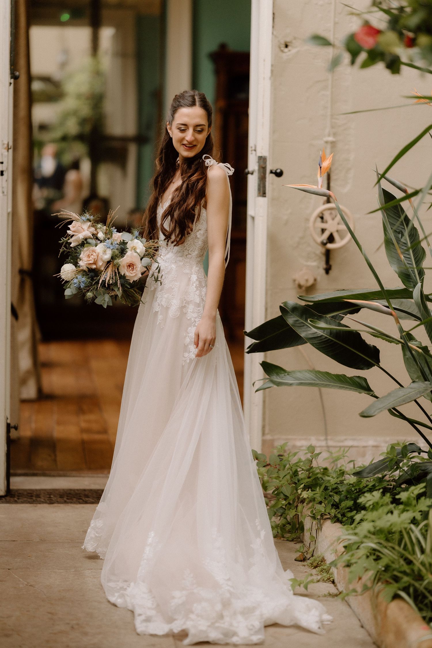 A bride in a flowing white lace gown holds a romantic blush and white floral bouquet while standing in a doorway at Prestwold Hall.
