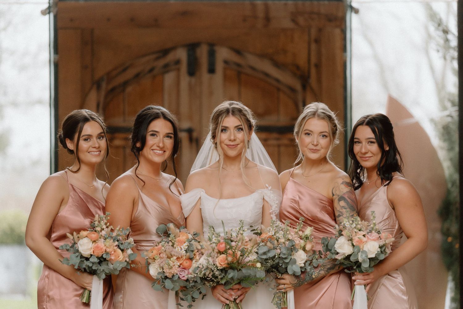 Wedding party holding peach and blush bouquets in front of a rustic venue entrance at Shustoke Barn.