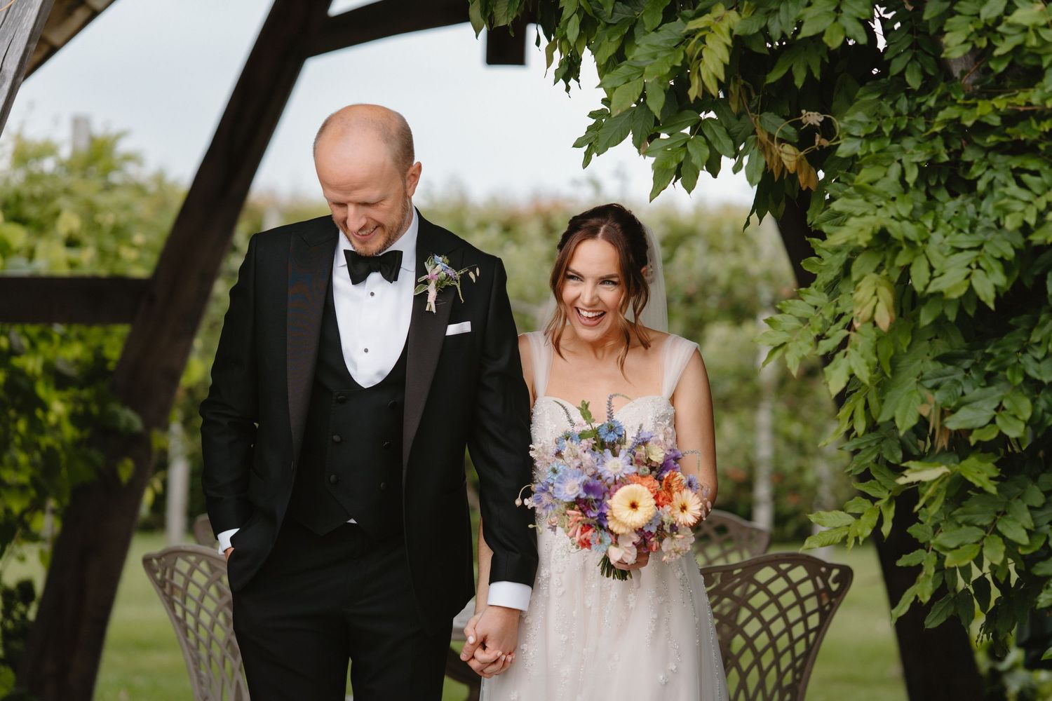 Bride and groom walking hand in hand through lush garden arbor, bride holding colorful bouquet in elegant wedding attire.