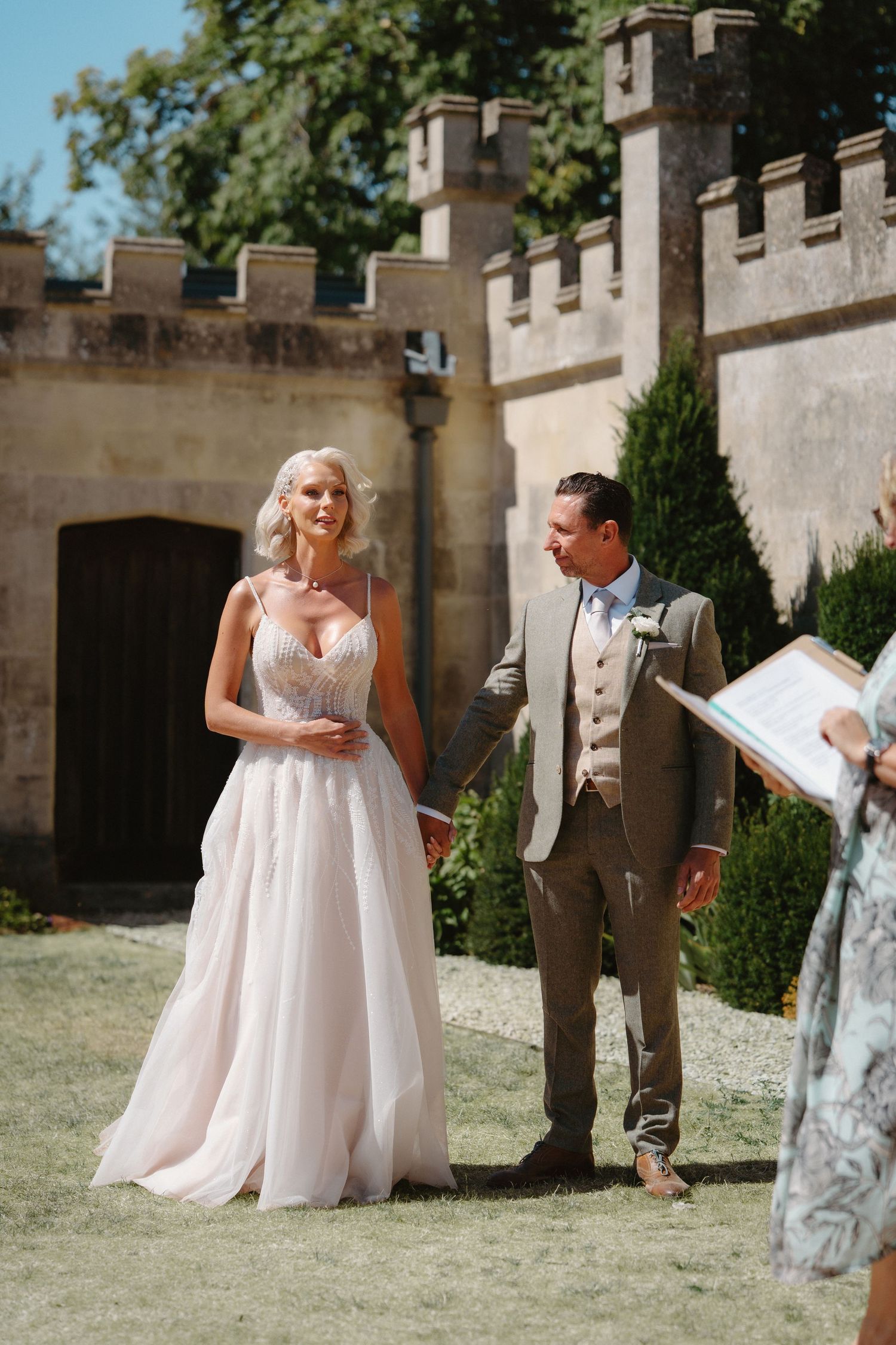 Bride in white gown and groom in tan suit stand together outdoors at a castle wedding ceremony.