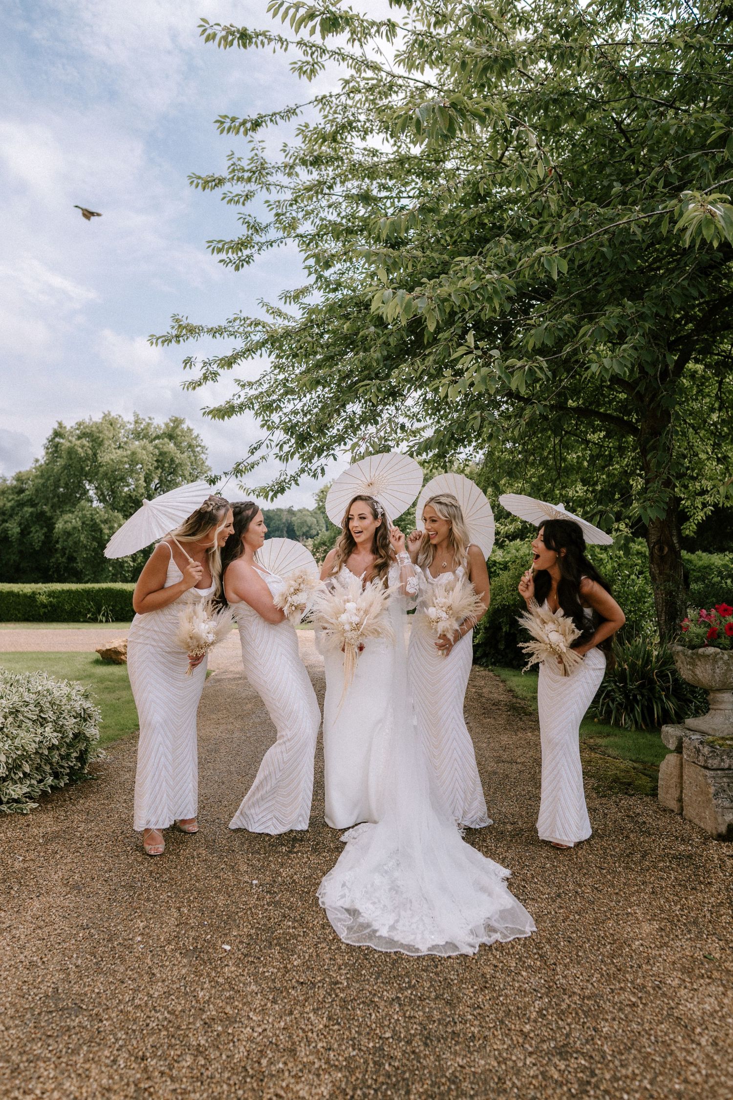 A bridal party in white dresses poses with parasols. Wedding photography at Rushton Hall. Northamptonshire Wedding photographer.