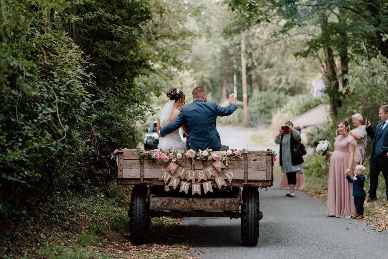 A rustic wagon decorated with wooden logs drives down a tree-lined road as wedding guests in pastel dresses look on.