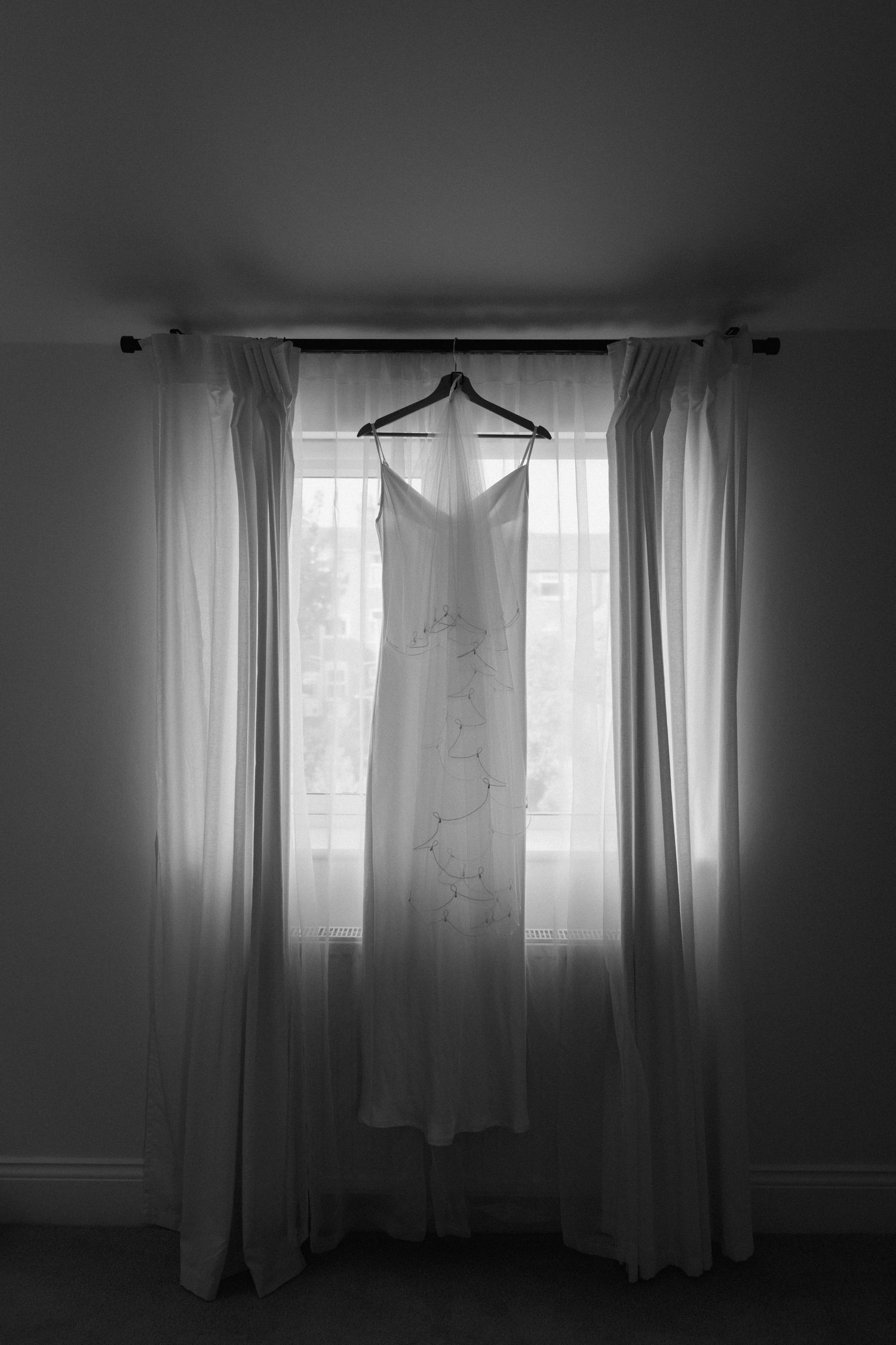 A white wedding dress hangs in silhouette against sheer curtains in a window.