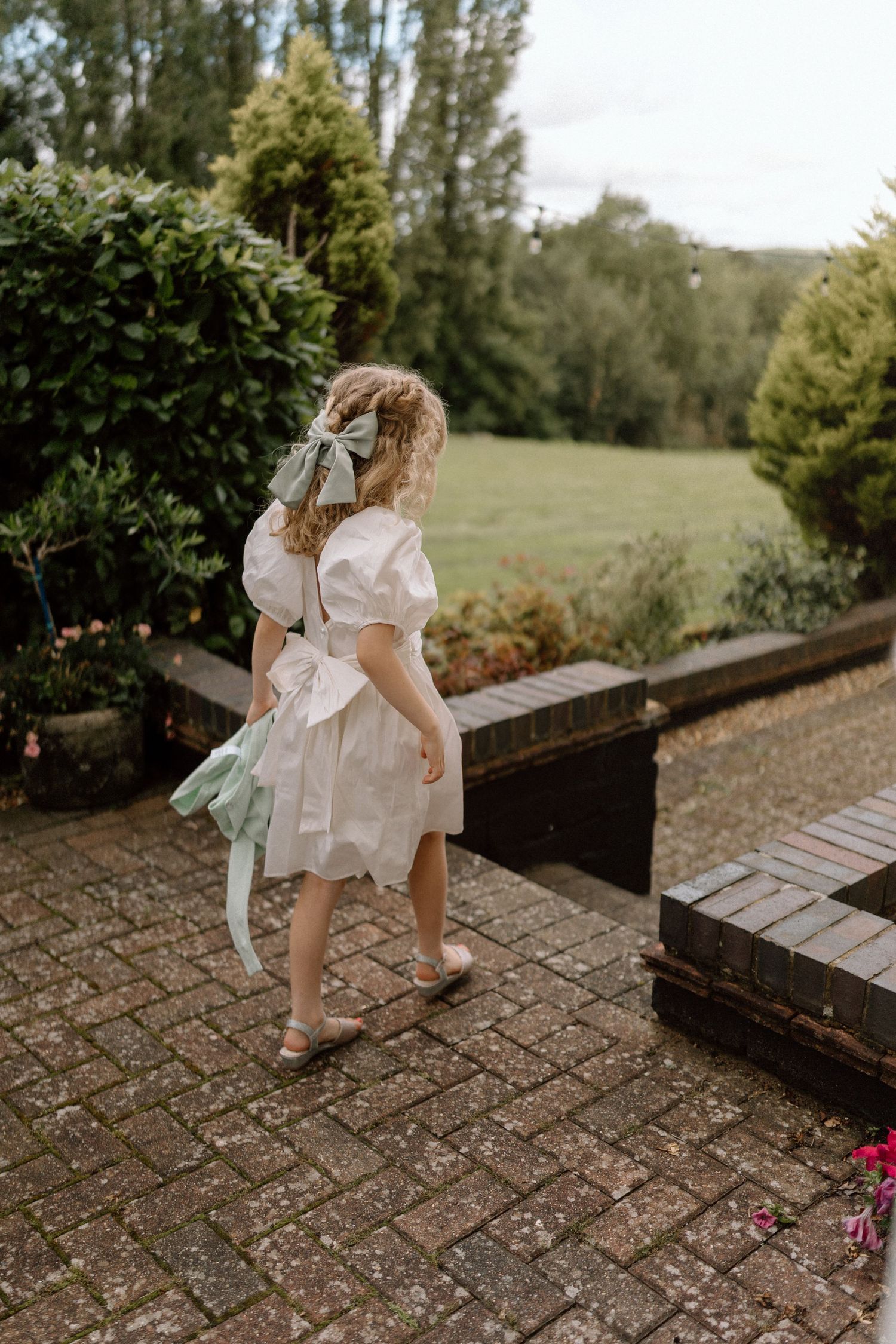 Young attendee in white dress walking along garden path during outdoor wedding.