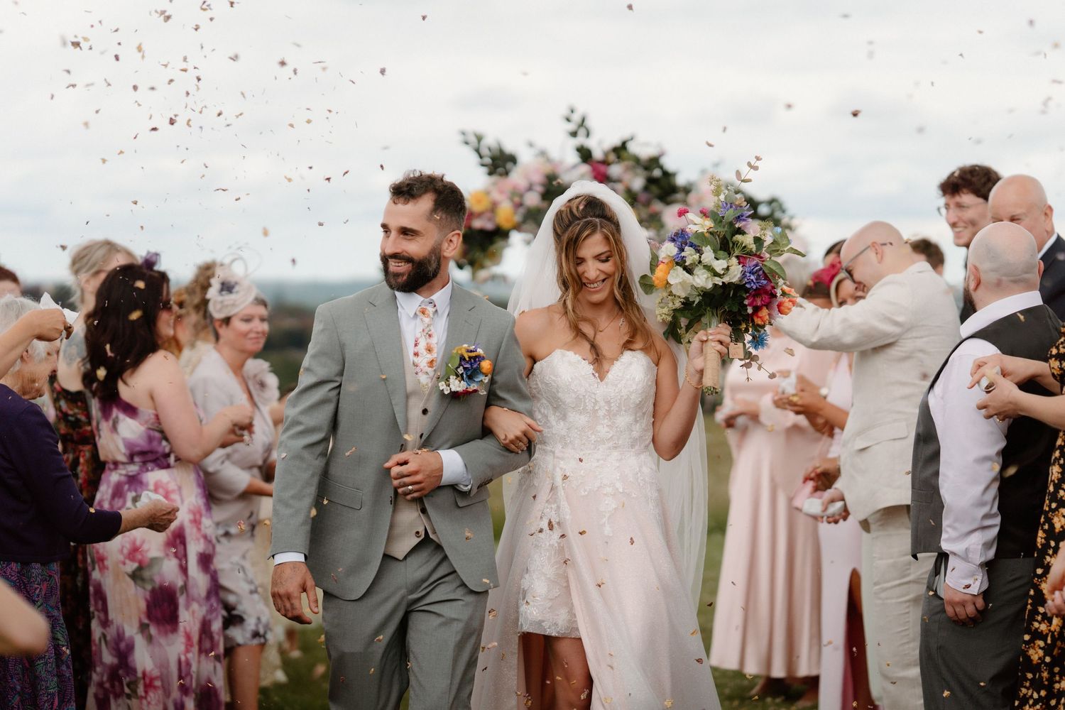 Bride and groom walk through confetti shower outdoors, guests celebrating joyful wedding ceremony on green hillside.