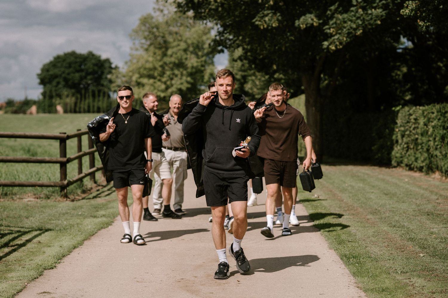 A sequence of people walking or jogging on a grassy path under sunlit trees.