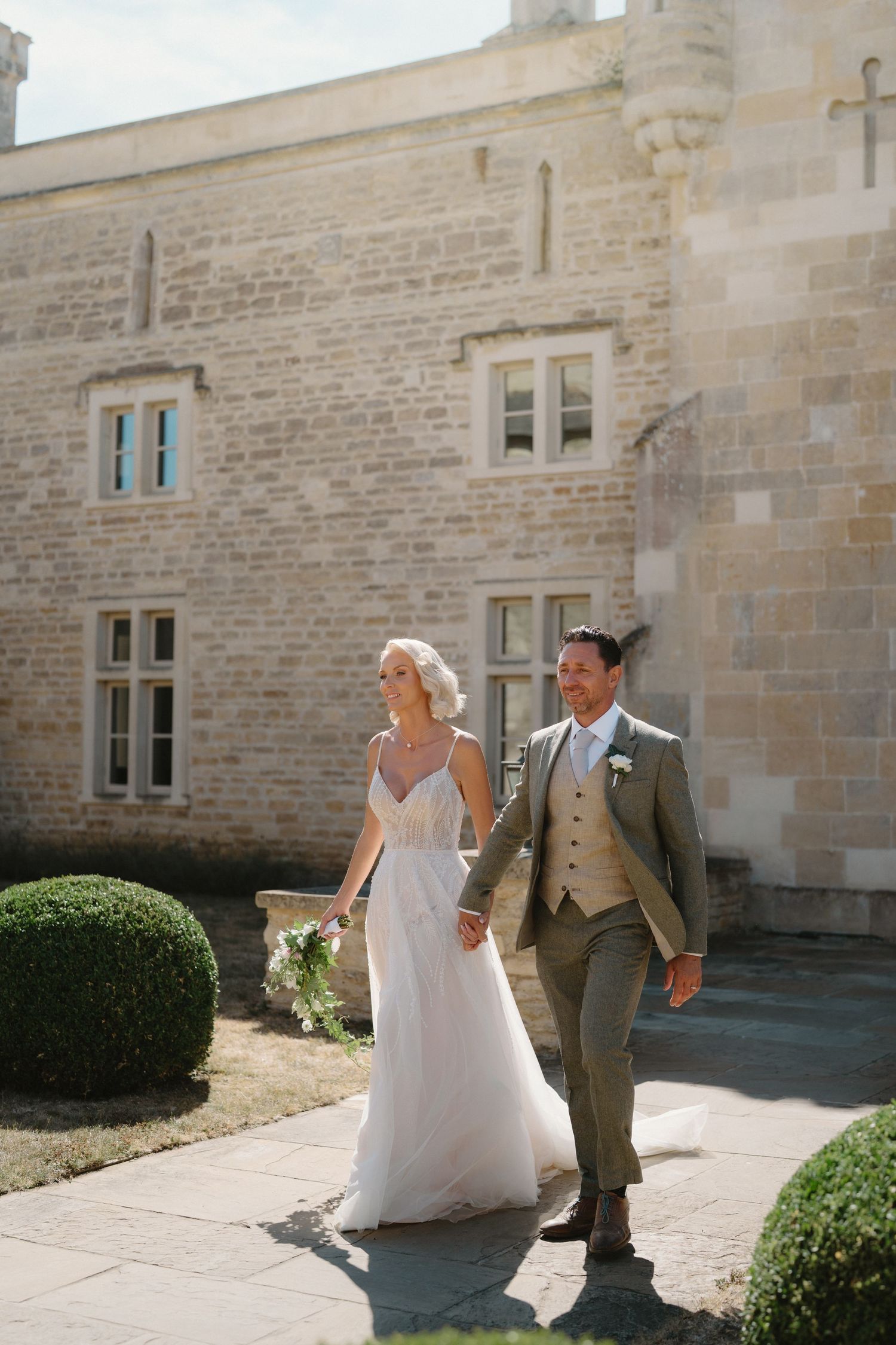 Bride and groom walk hand in hand along a sunlit stone path beside a historic castle wedding venue.