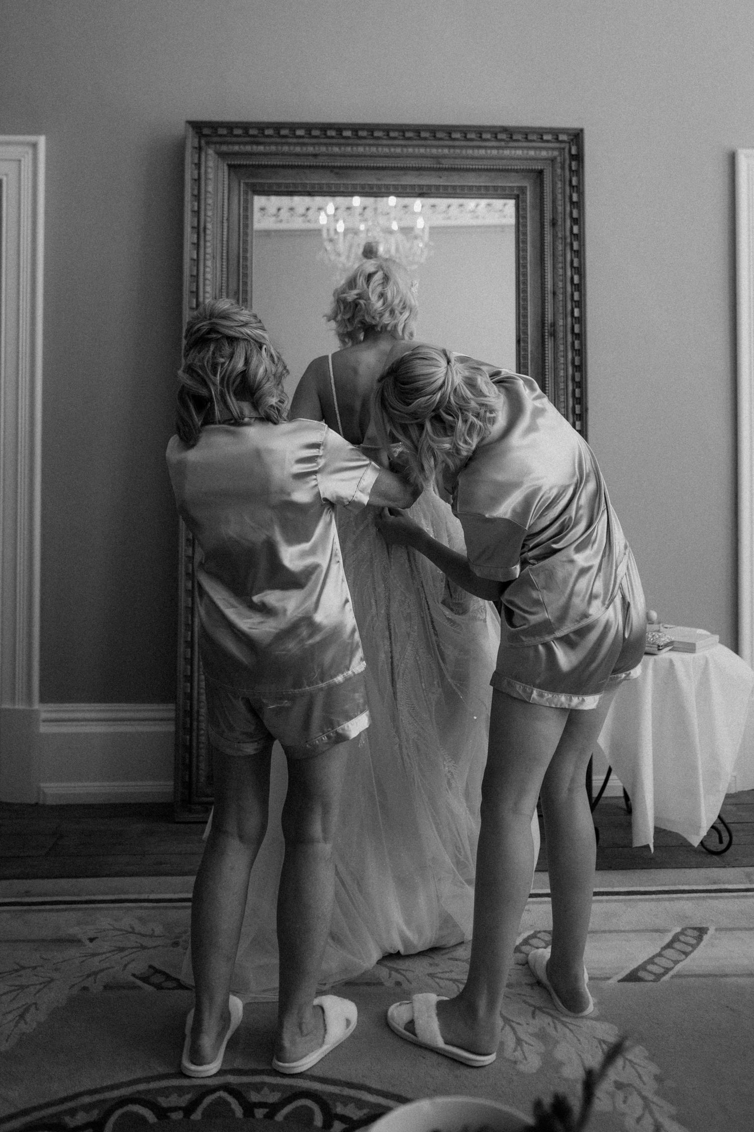 Bridesmaids in satin robes help bride with wedding dress in front of ornate mirror, black and white photo.