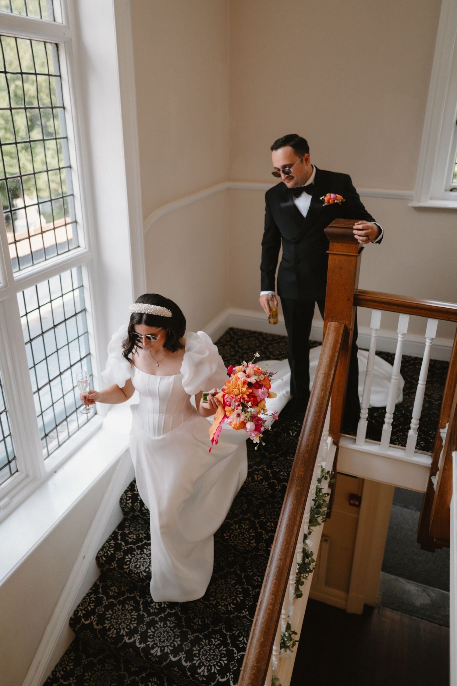 Bride in white gown holding colorful bouquet descends staircase with groom in black tuxedo at elegant wedding venue.