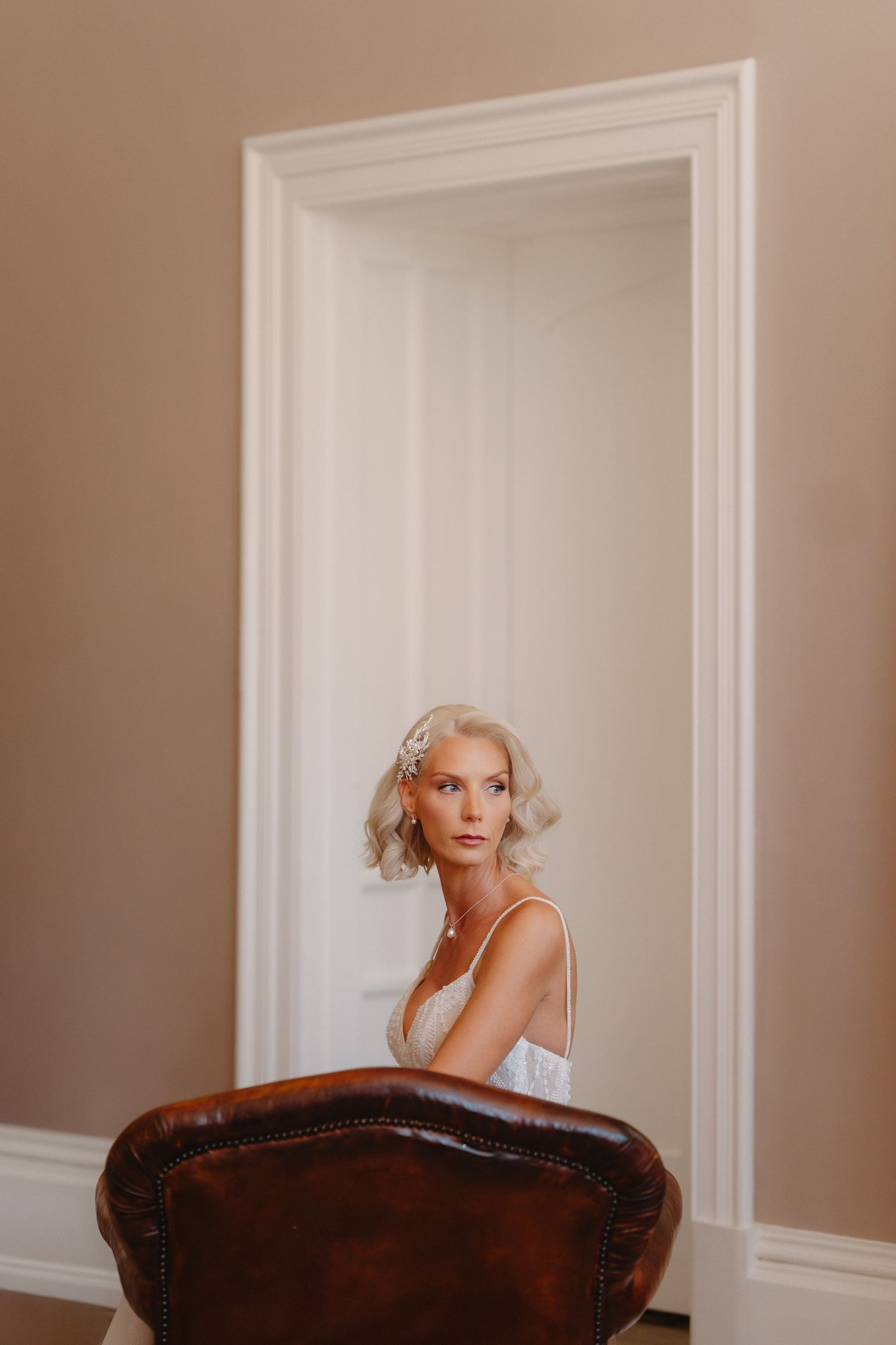 Elegant blonde bride in white lace gown posing in a doorway with a vintage leather chair in a beige room.