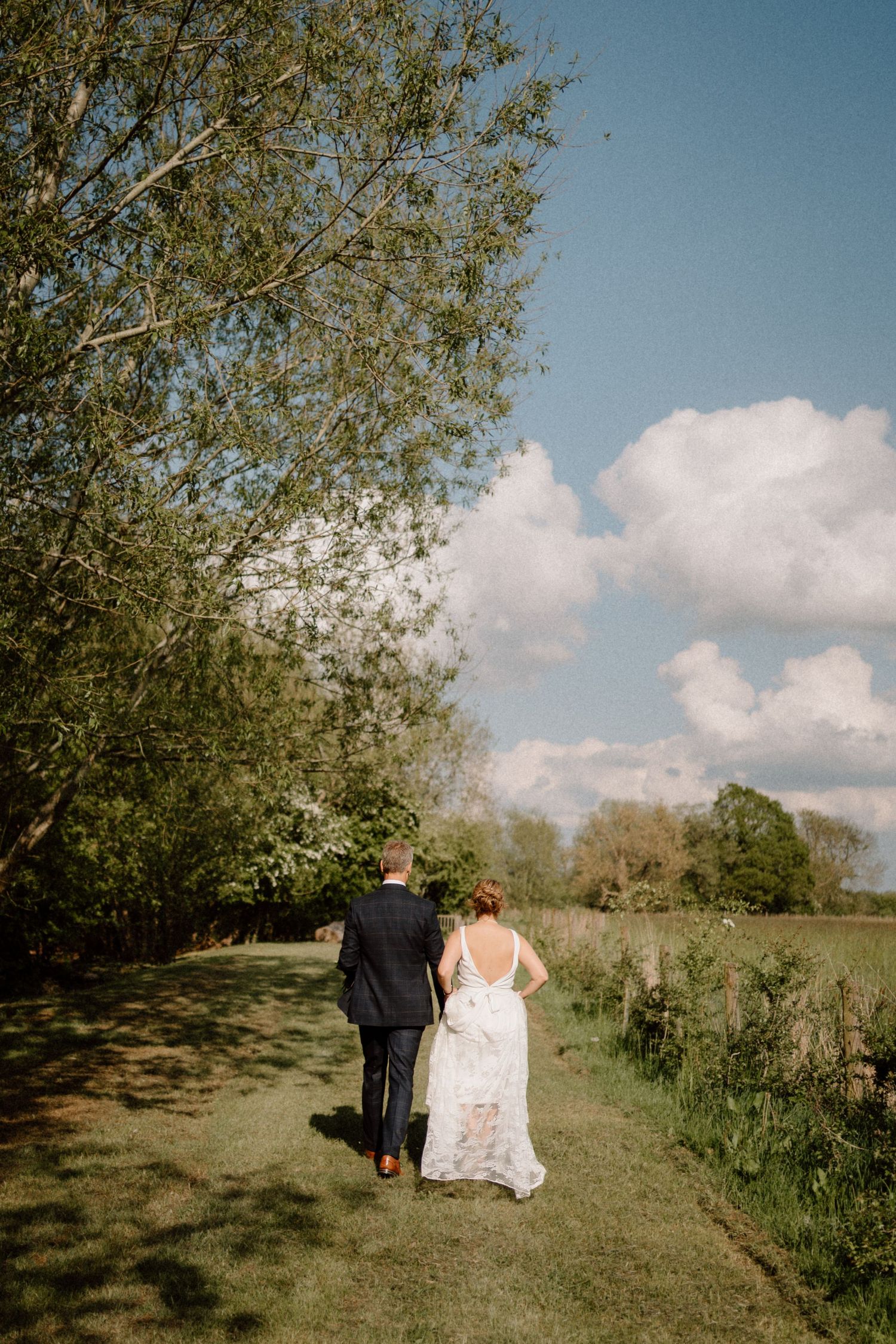 A couple in wedding attire walk down a tranquil nature path with trees and white clouds in a blue sky overhead at Swallows Nest Barn.