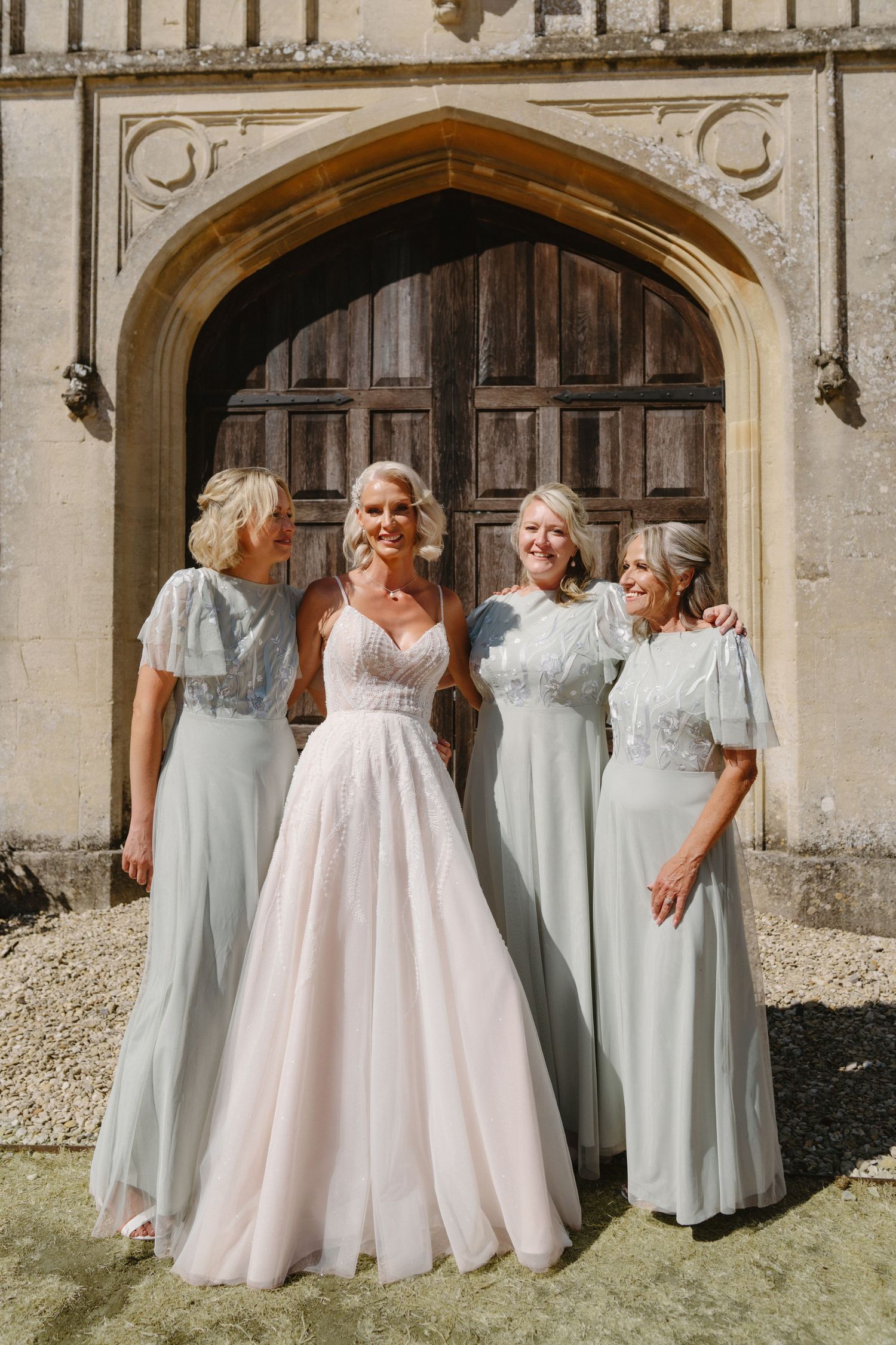 Bride in blush gown with three bridesmaids in sage dresses outside gothic stone arch.
