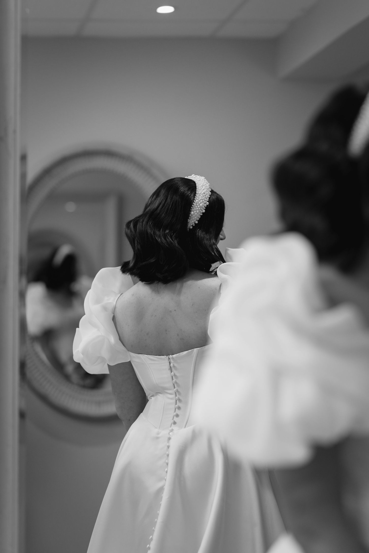 Bride in white off-shoulder wedding gown with pearl headband viewed from behind in mirror reflection.