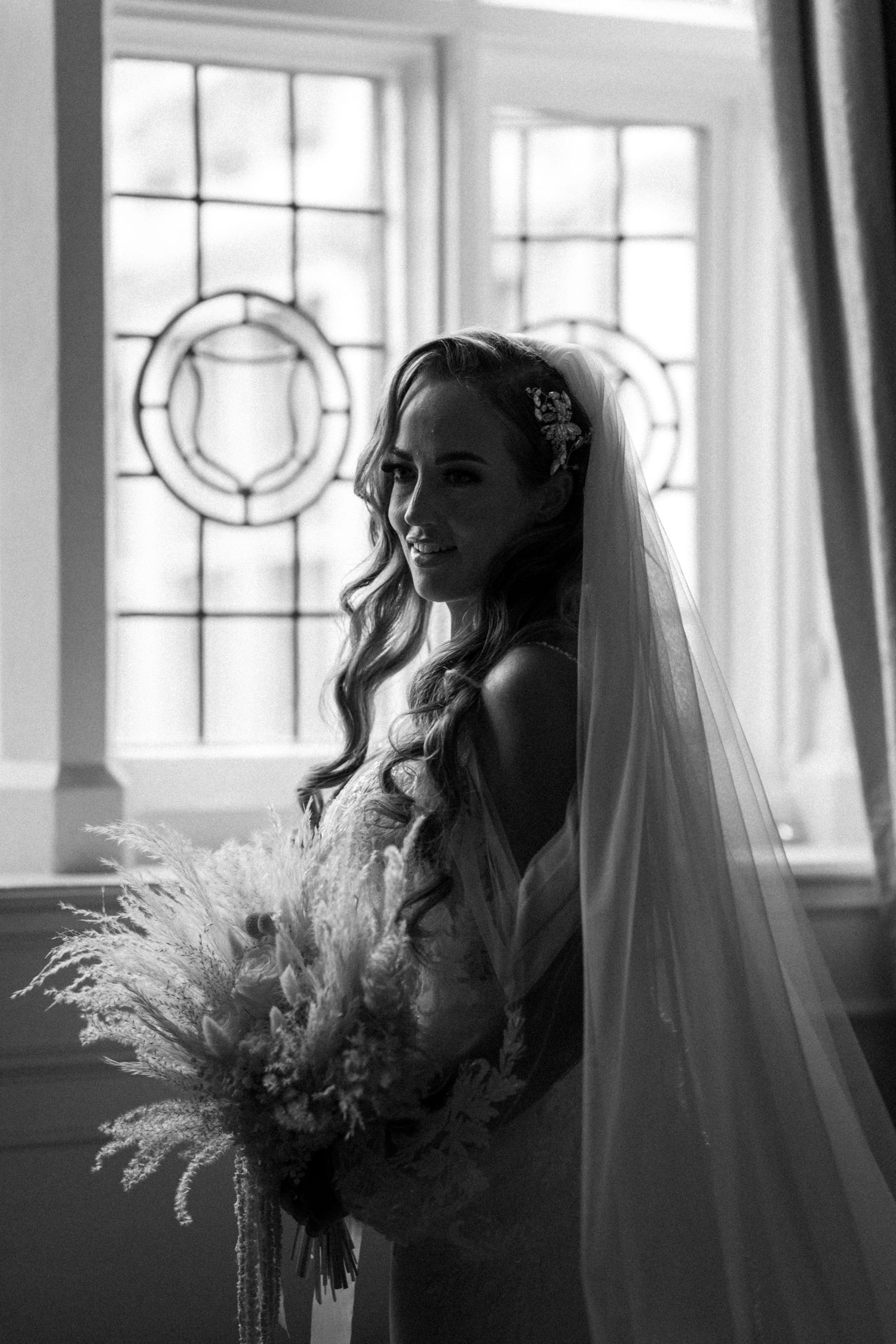 A black and white photo series showing a bride with a flowing veil holding a feathered bouquet in various poses near windows.