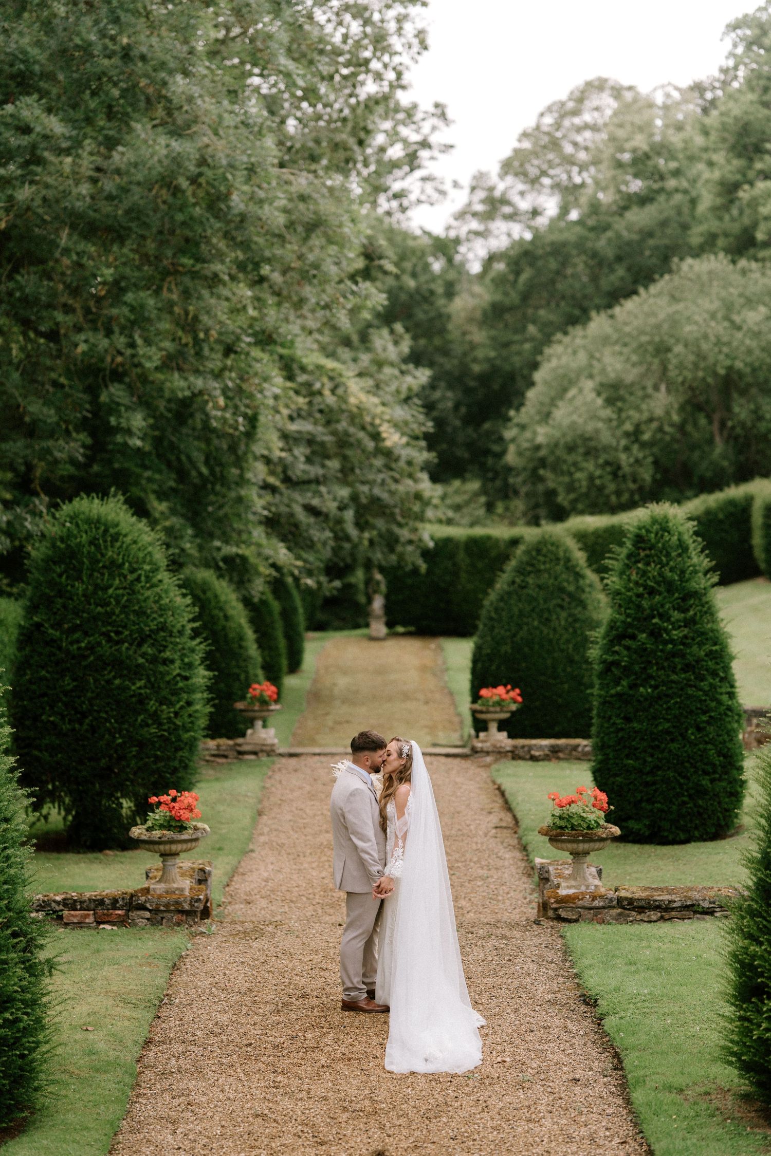 A wedding couple embraces on a garden path lined with perfectly manicured topiary and colorful flowers.