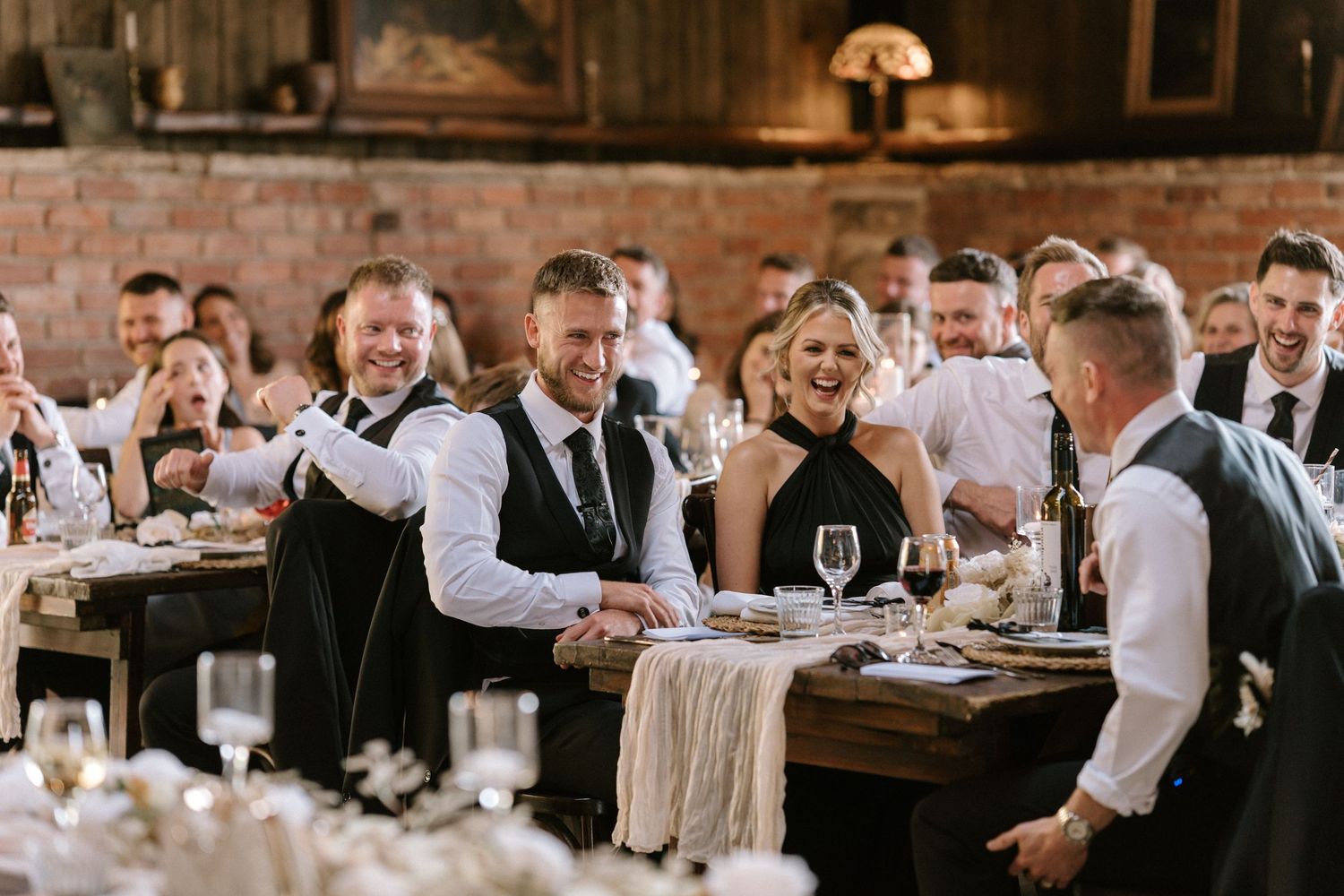 Wedding reception dinner scene in rustic venue with guests seated at long wooden tables.