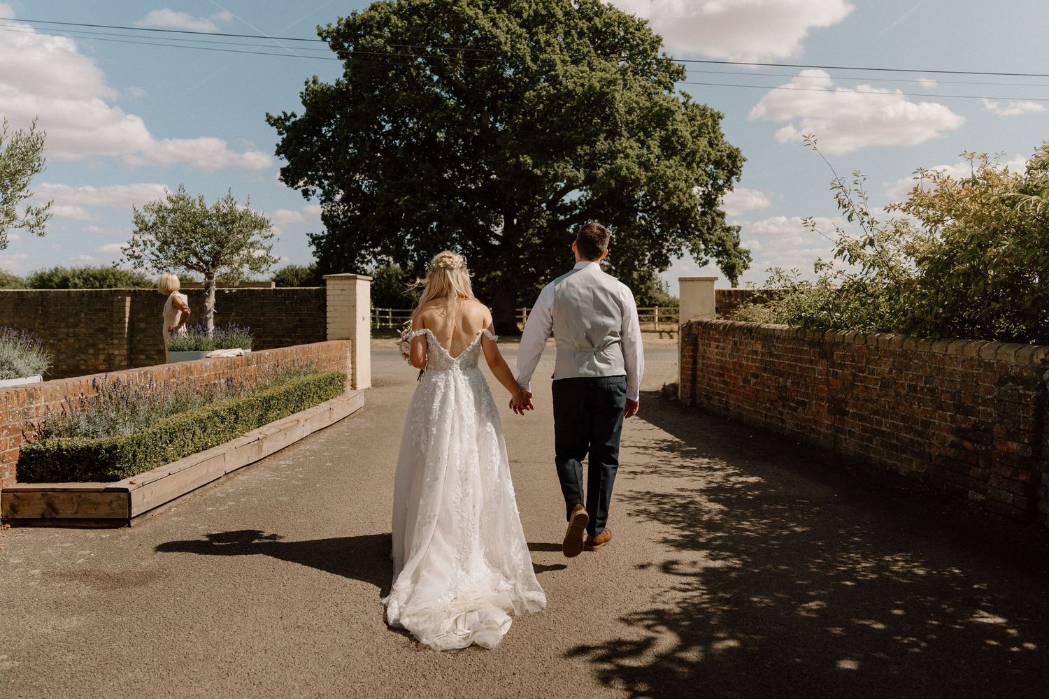 A couple walking away down a sunny path with stone walls, the bride wearing a flowing white lace wedding dress at Milling Barn.