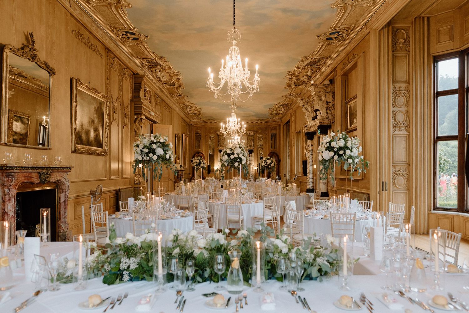 An elegant reception room with ornate ceiling, crystal chandeliers and white floral table settings at Harlaxton Manor.