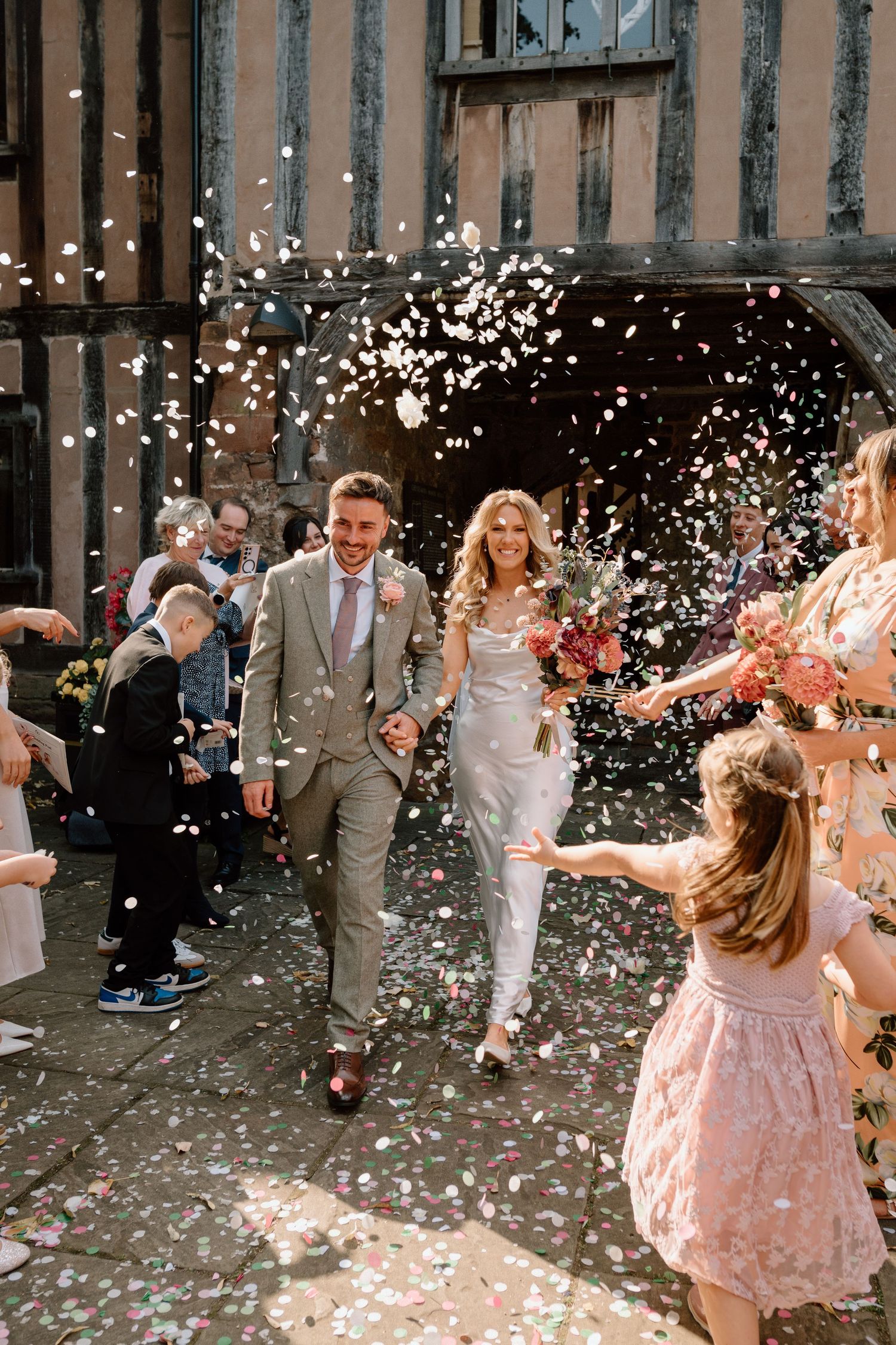 Newlyweds walk through a shower of confetti while guests celebrate at a rustic venue at Cheylesmore Manor in Coventry.