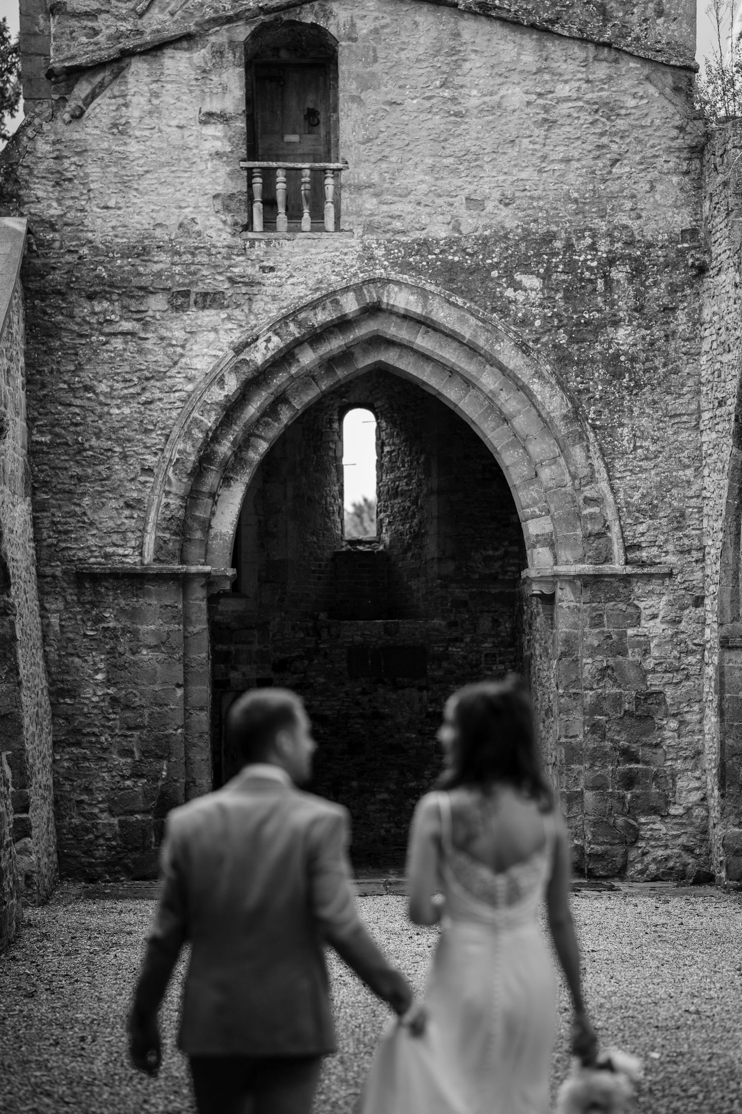 A couple in wedding attire viewed from behind approaching a historic stone archway entrance at Ettington Park Hotel.
