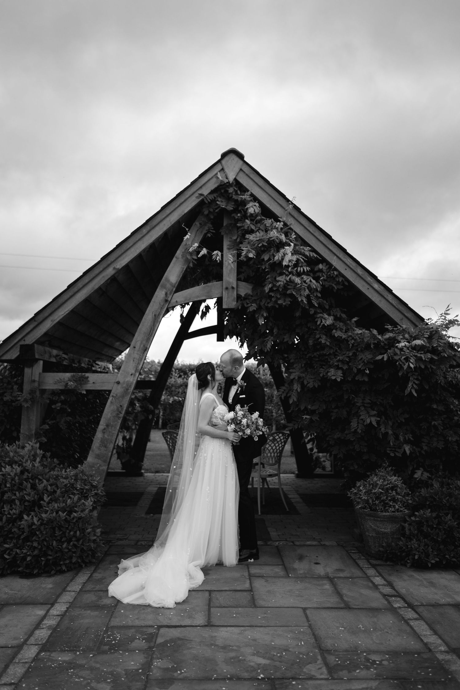 Black and white photo of bride and groom kissing under a wooden pergola arch surrounded by lush greenery on their wedding day.
