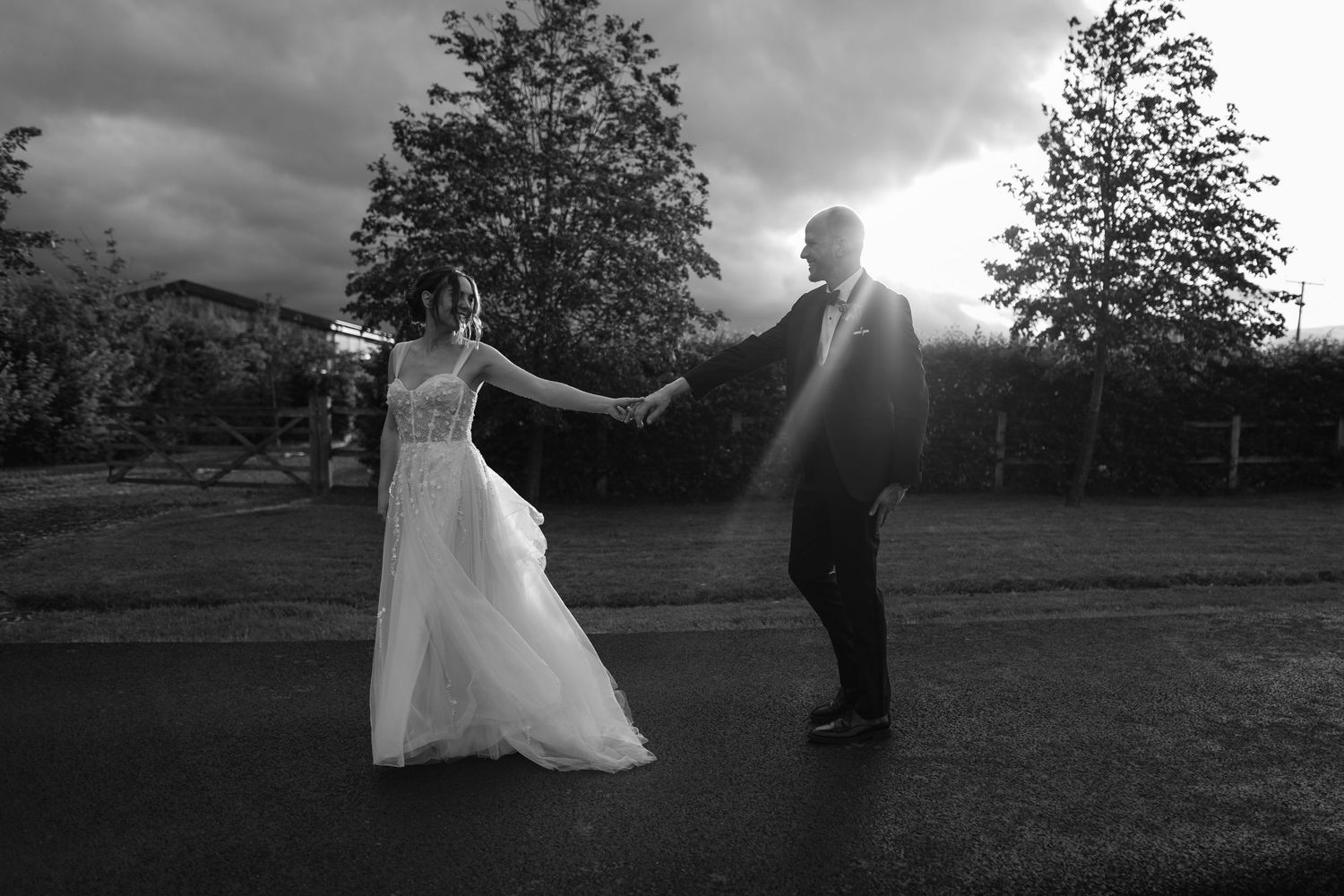 Black and white photo of a bride and groom dancing outdoors at dusk, holding hands with trees in the background.
