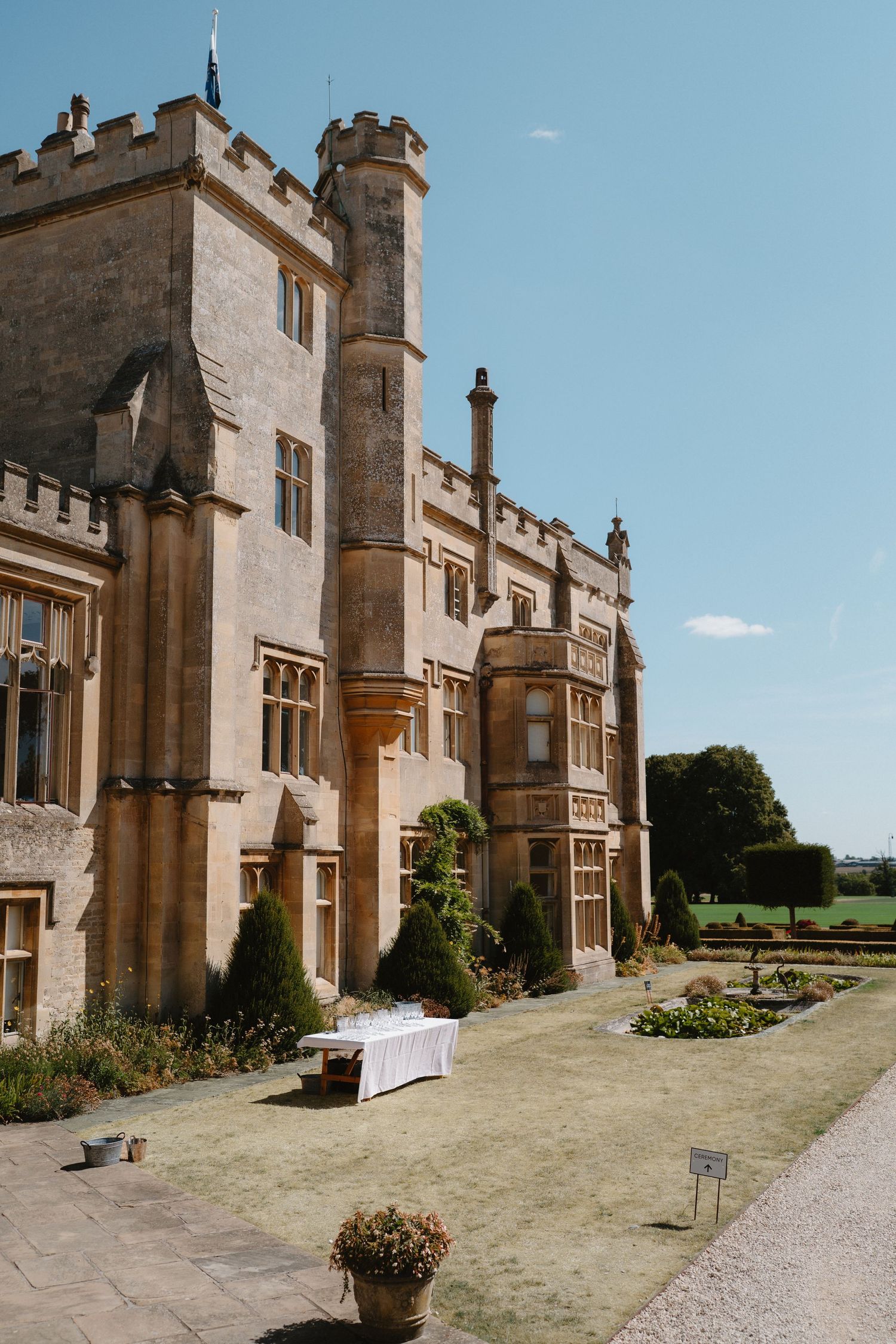 Historic English manor house with Gothic architecture, manicured gardens, and a white-clothed table on the lawn under blue sky.