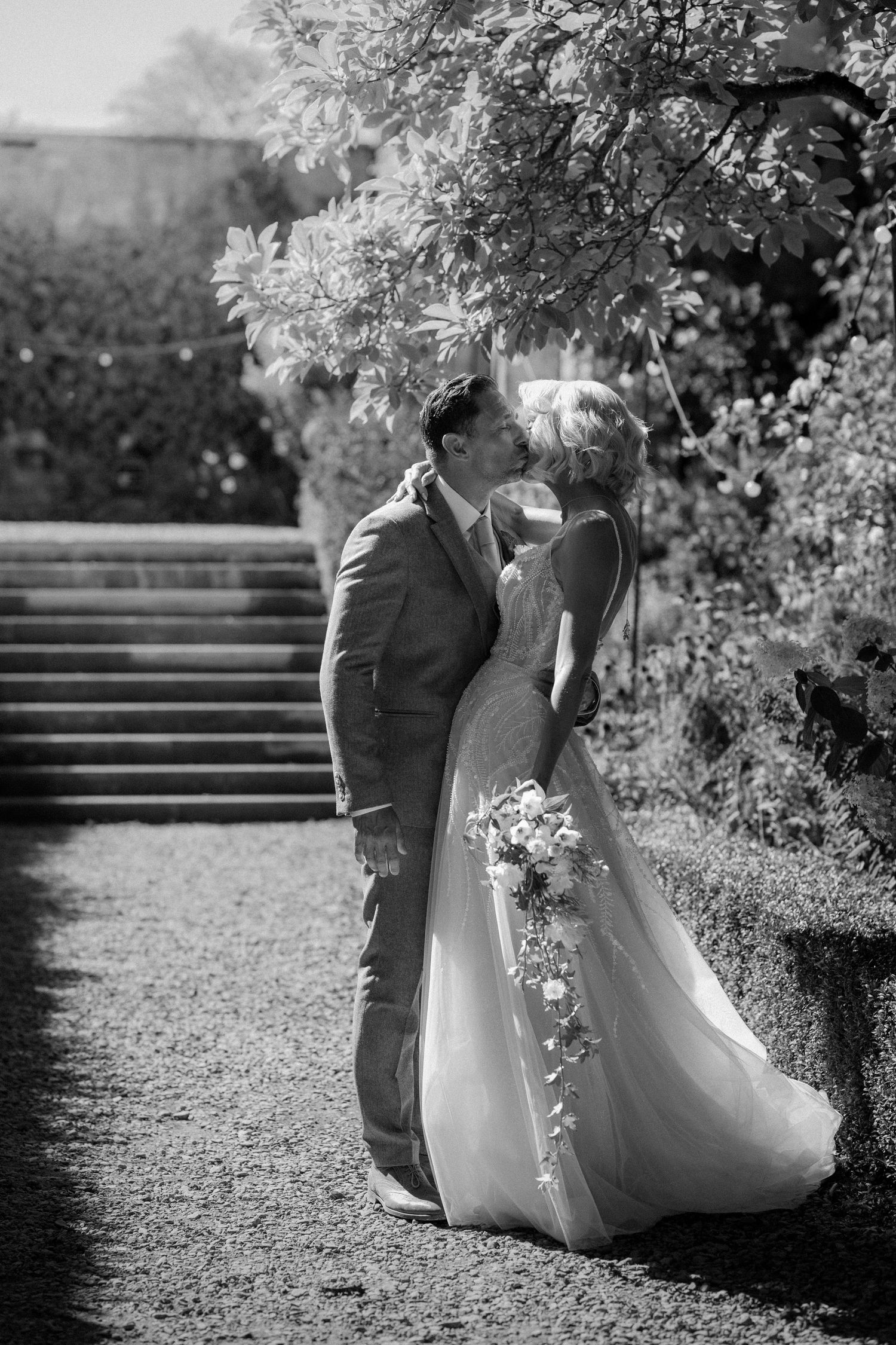 Newlyweds share a romantic kiss in a lush garden, bride holding a cascading floral bouquet.