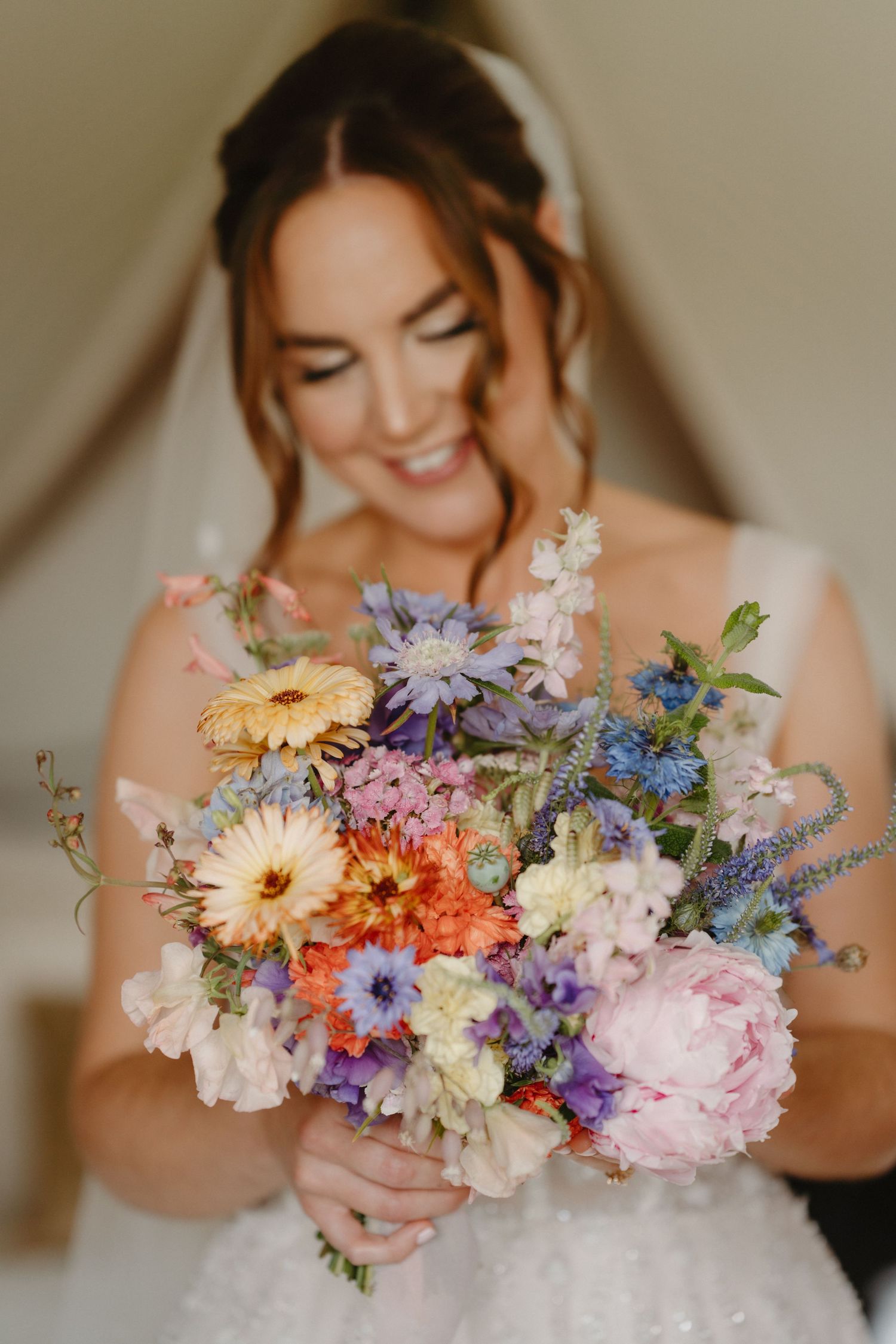 Smiling bride in white gown holds a colorful wildflower bouquet with peonies, gerberas, and lavender blooms.
