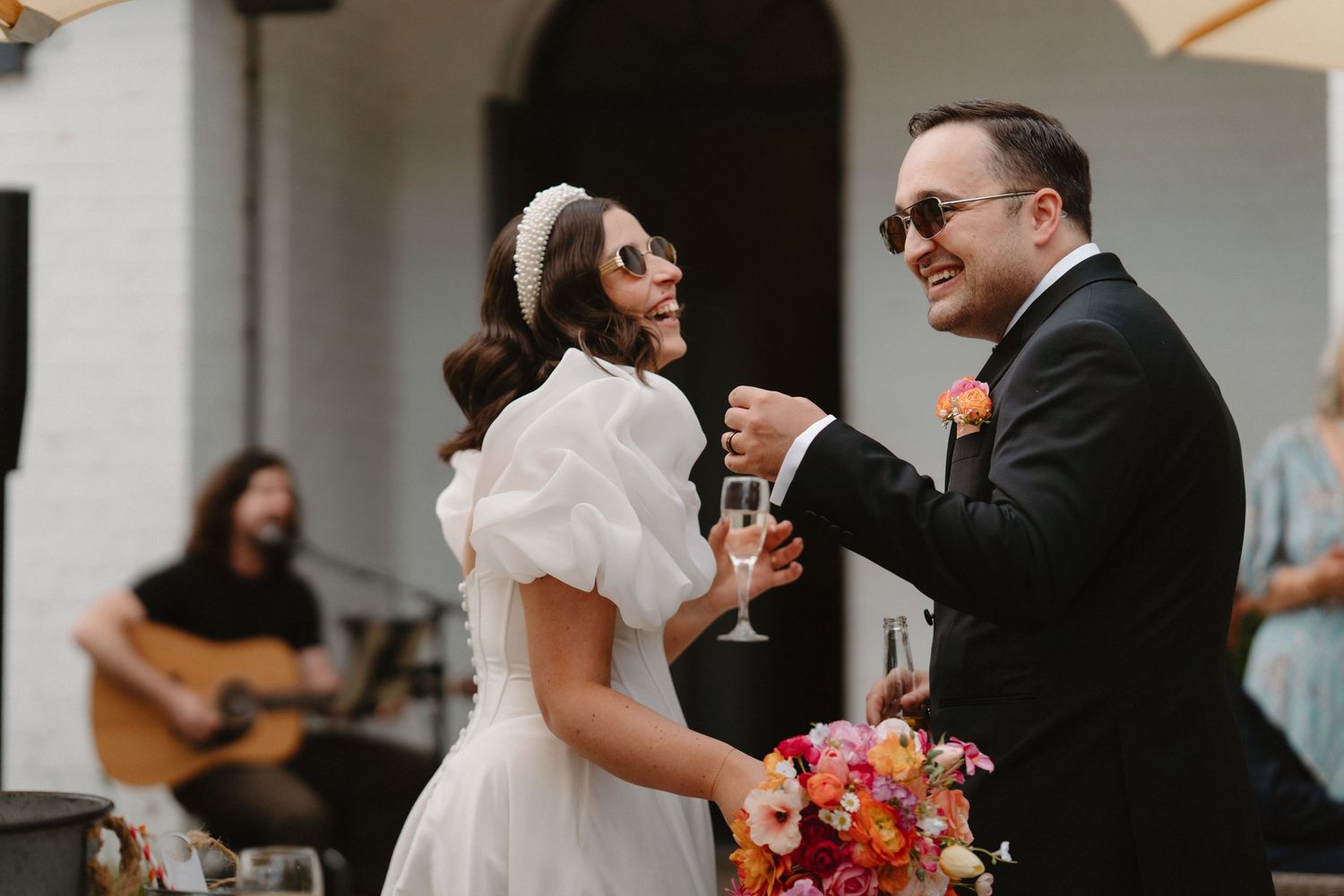 Bride and groom toasting with champagne glasses at wedding reception with guitarist performing in background.